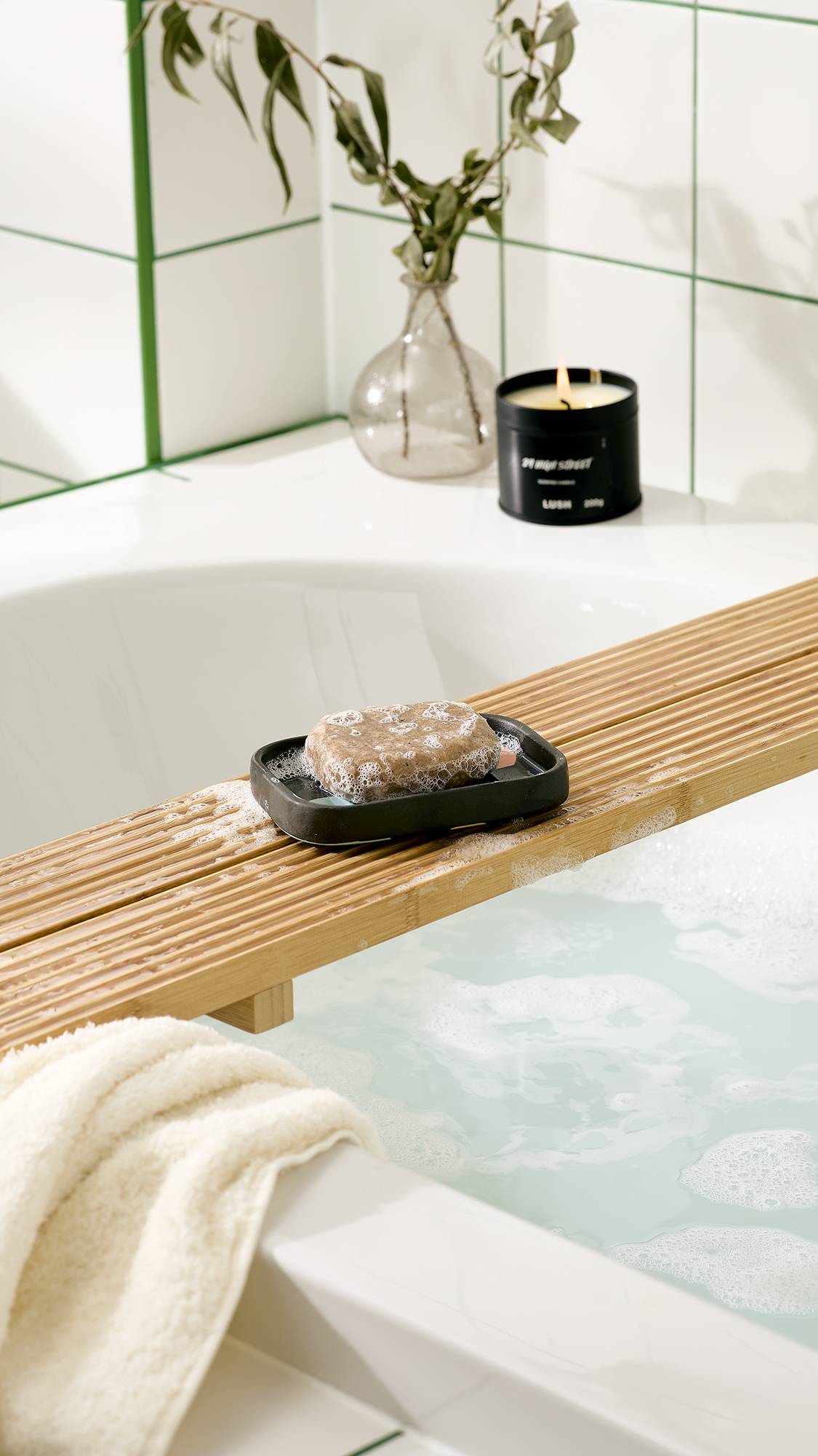 A bar of bubbly soap rests on a dark grey ceramic soap dish with pastel spots, set on a wooden bath caddy over foamy bathwater. A black candle, vase, and white towel sit nearby against white tiled walls.