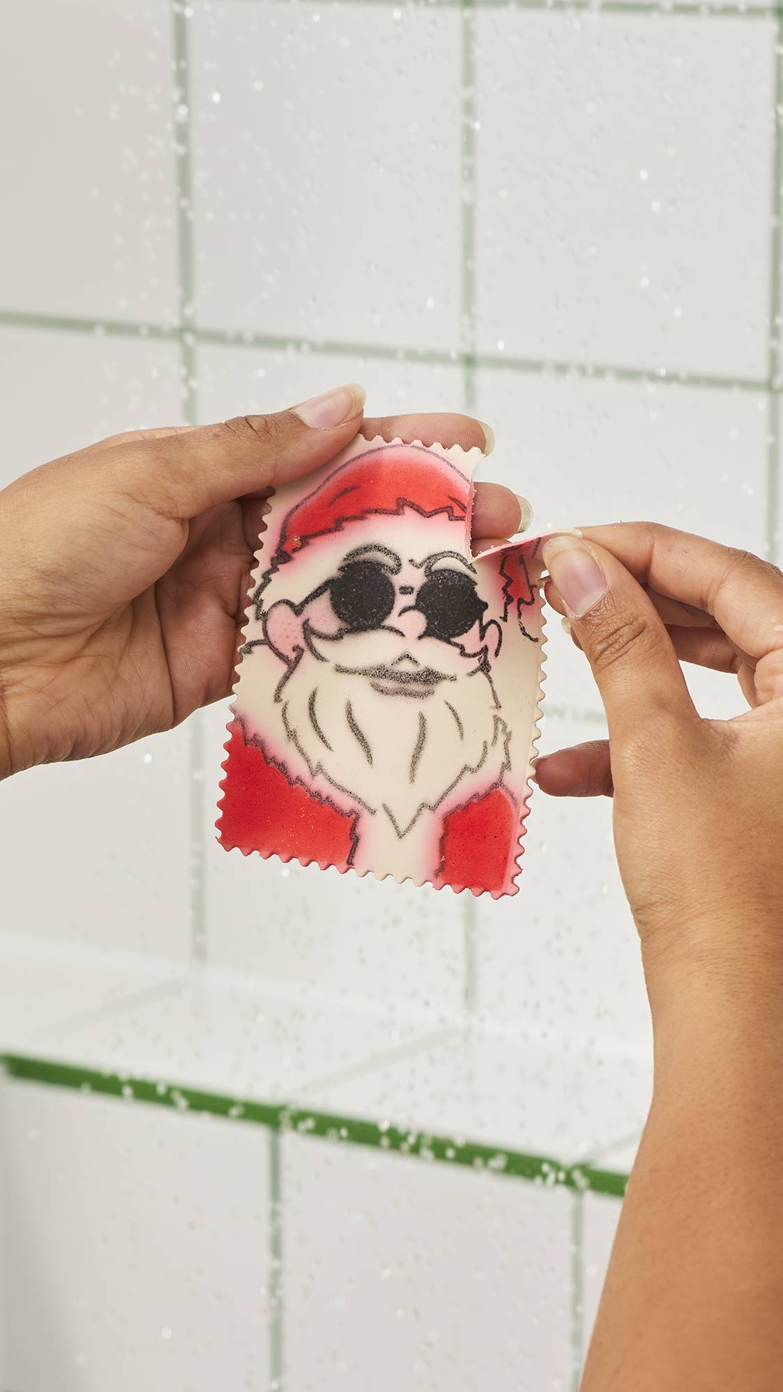 Hands tear off a single strip from the side of a surfactant paper sheet shaped like a postage stamp featuring Santa Claus wearing sunglasses, in the shower against a white tiled wall background.