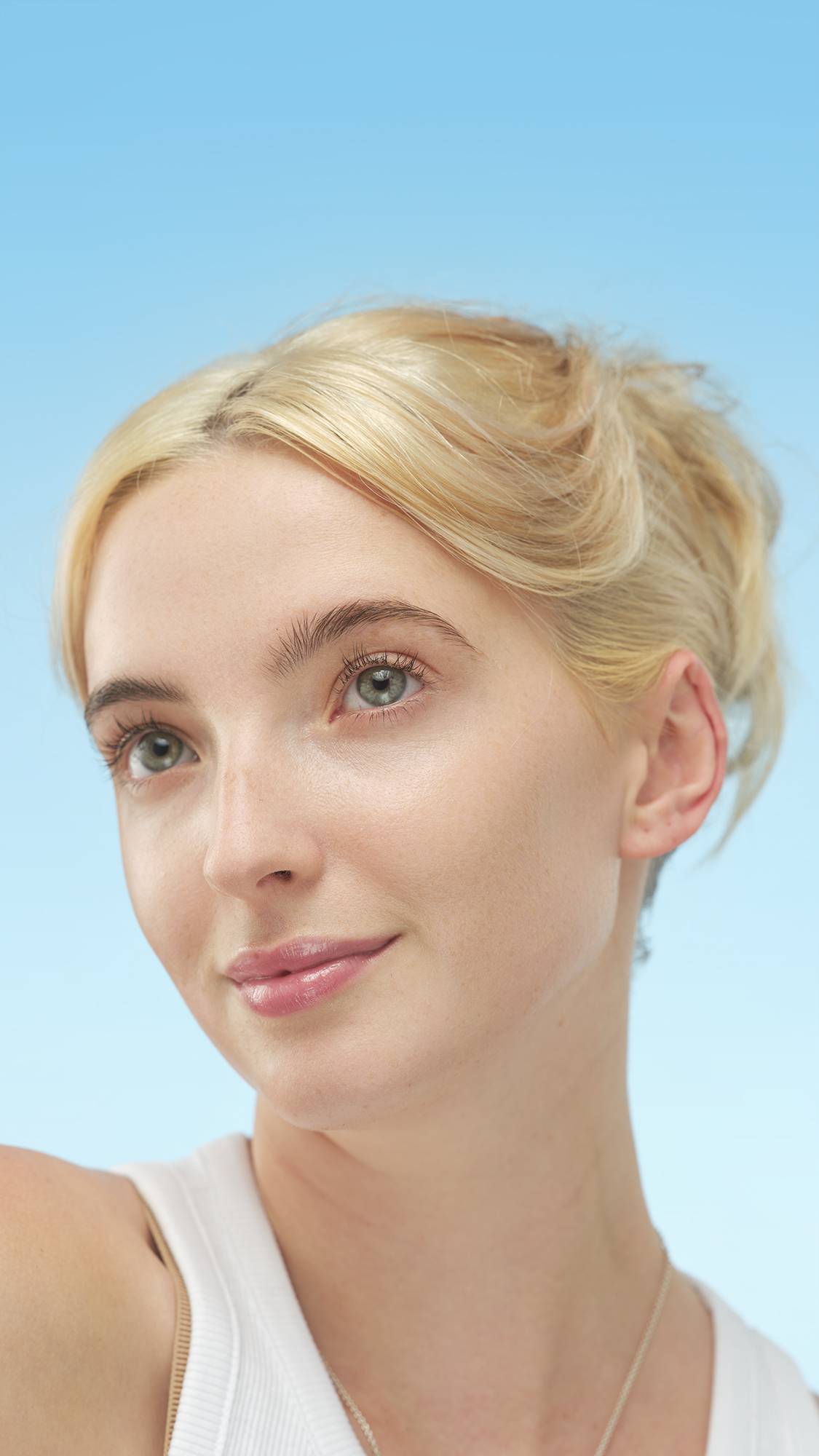 A close-up vertical portrait of a person with fair skin, light blonde hair, and light eyes, wearing Light Cool Skinwear. They look up and right, set against a bright, gradient blue background.