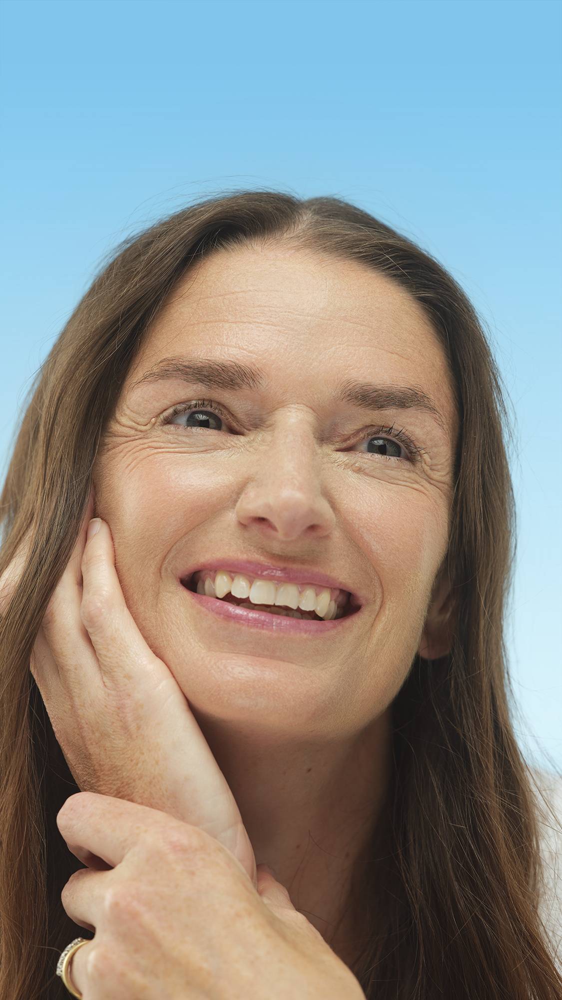 A close-up vertical portrait of a smiling person with warm-toned skin and long brown hair, looking up. Their hand rests on their cheek, and their face has a warm glow from the Light Medium Warm Beauty Balm, set against a bright blue background.