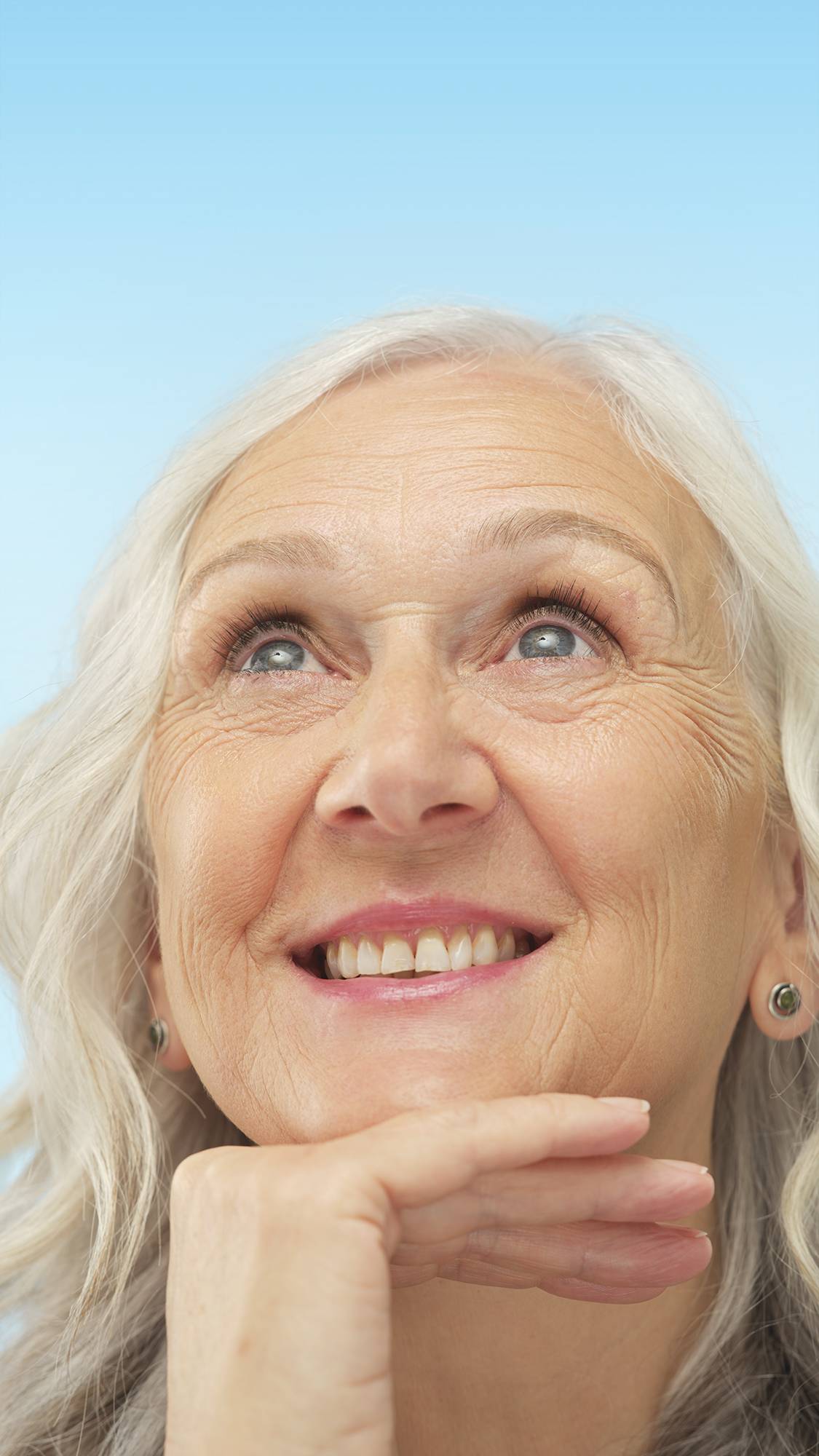 A smiling person with silver hair and Light Neutral Skinwear foundation applied, looks upward with their chin resting on their hand against a light blue background.