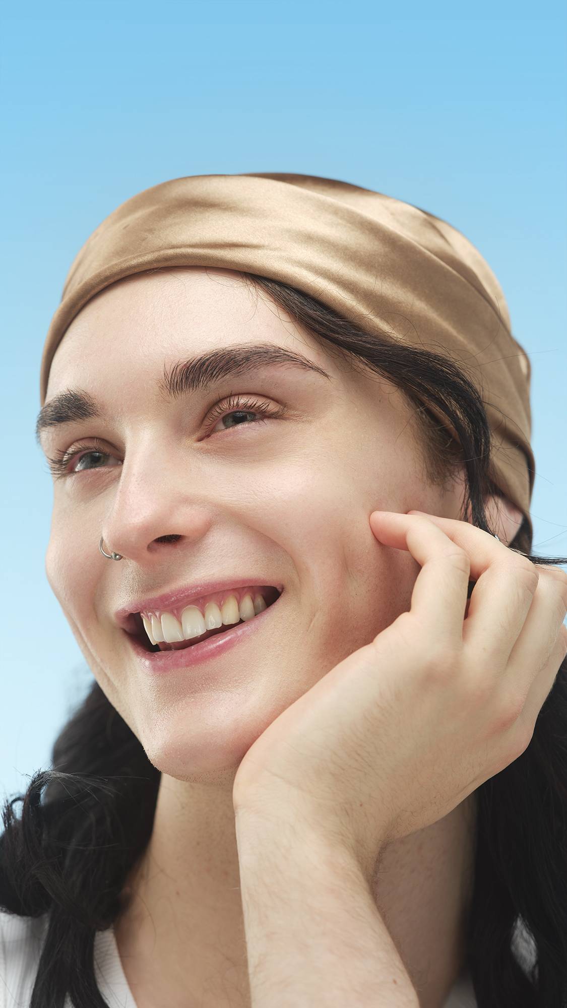 A close-up vertical portrait of a smiling person wearing a silky, golden-tan headband. Their skin is enhanced with the Light Warm Skinwear, and one hand rests on their jaw, all set against a bright blue sky.