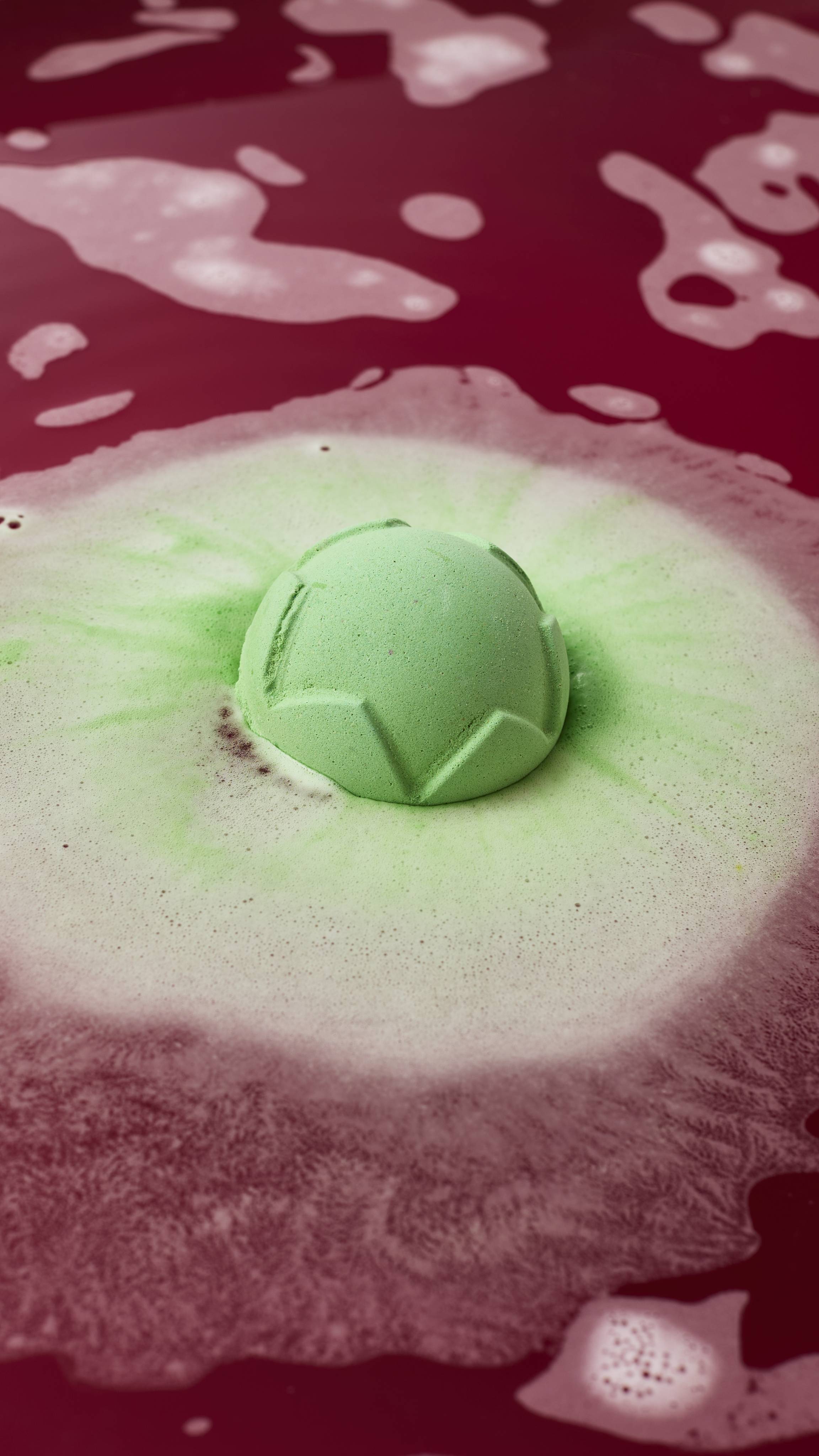 A carved light-green bath bomb floats in deep magenta bathwater, fizzing to create a spreading ring of pastel green and white foam around itself.