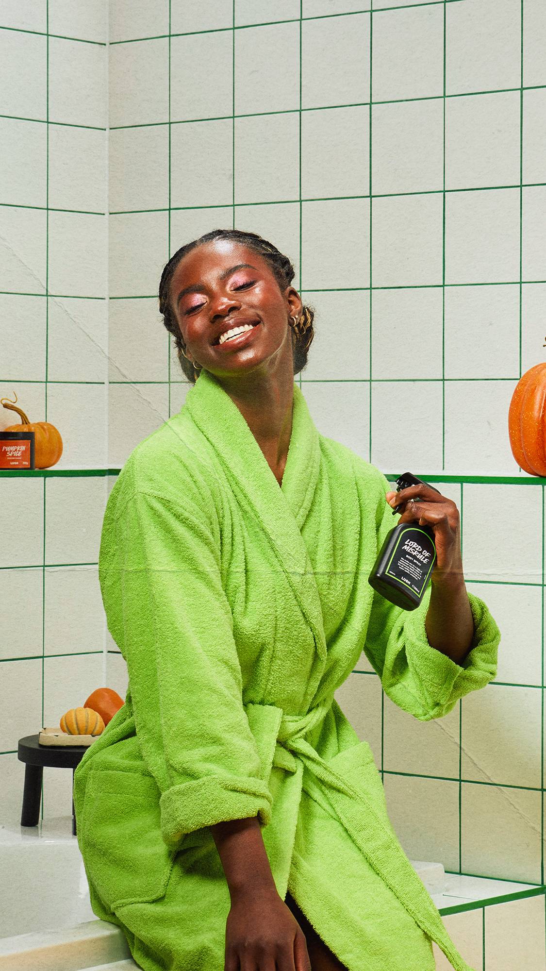 A smiling model in a bright green dressing gown holds a Lush Lord of Misrule body spray in a white-tiled bathroom with green grout, surrounded by seasonal pumpkins.