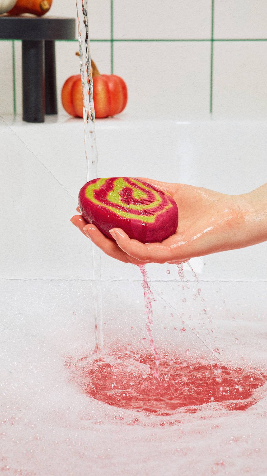 A hand holds a magenta and green "Lord of Misrule" bubble bar under running tap water, splashing vibrant pink water into a frothy, bubble-filled bath. A small orange pumpkin sits on the white-tiled tub edge in the background.