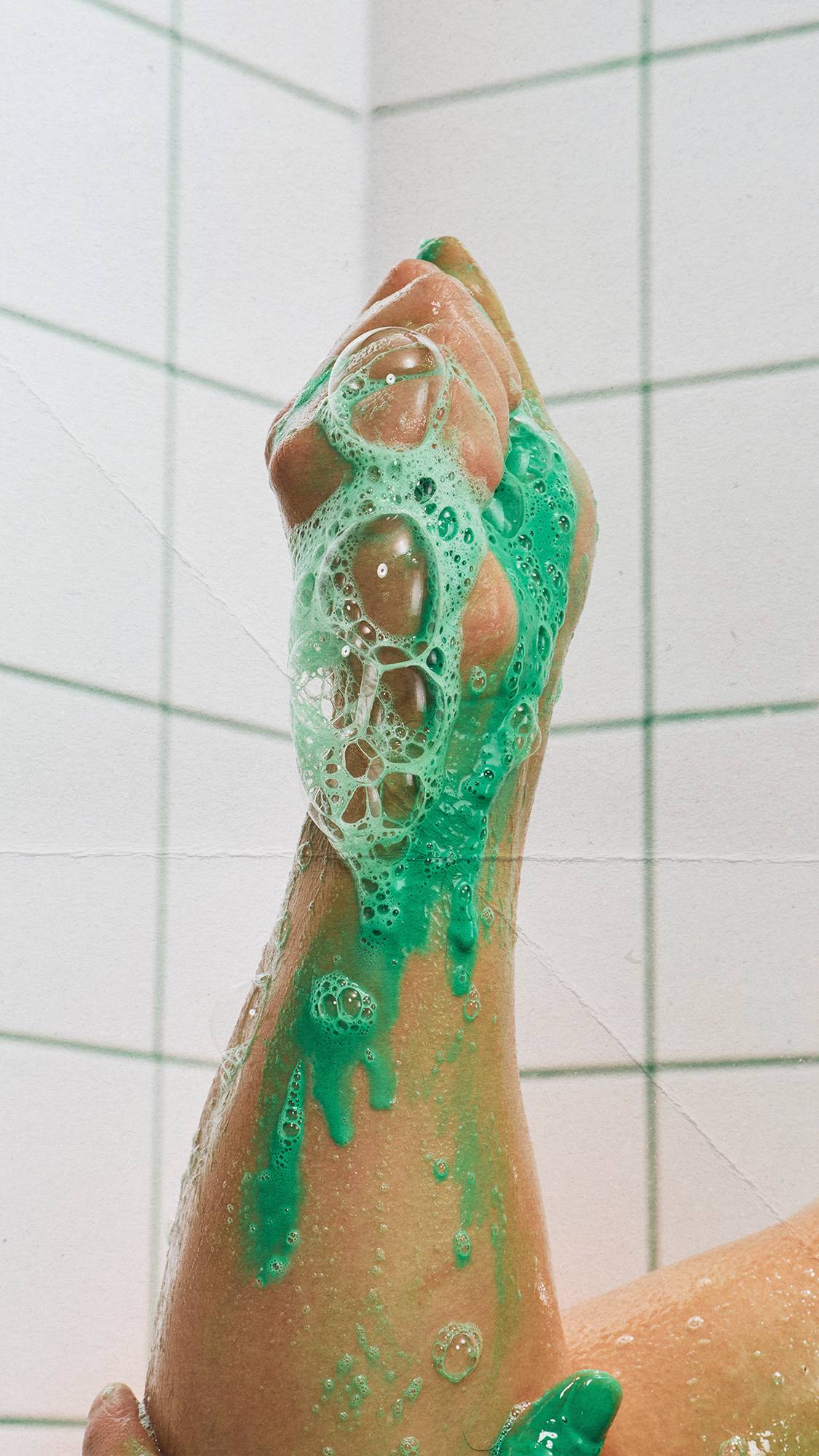 A raised, wet arm covered in bubbly, bright green "Lord of Misrule" shower gel lather, set against a corner backdrop of white wall tiles with green grout lines.