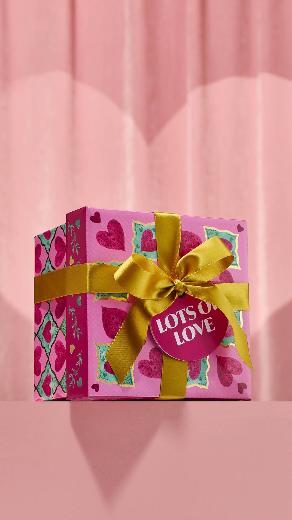 The Lots of Love Gift box features a pink design with hearts inside teal squares, tied with a thick gold ribbon. A round tag reading Lots of Love hangs from the bow against a soft pink curtain background.