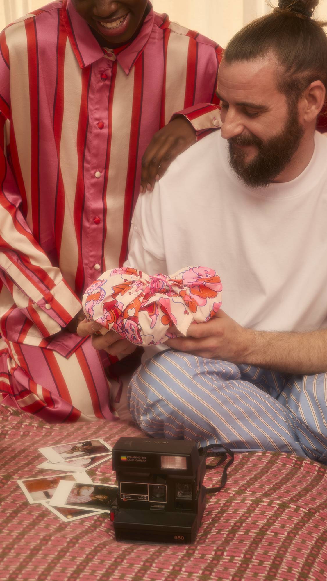 Two people sit on a patterned bedspread, with one holding a gift wrapped in the Love Bug Knot Wrap. An instant camera and scattered photos lie in the foreground as they smile at the package.