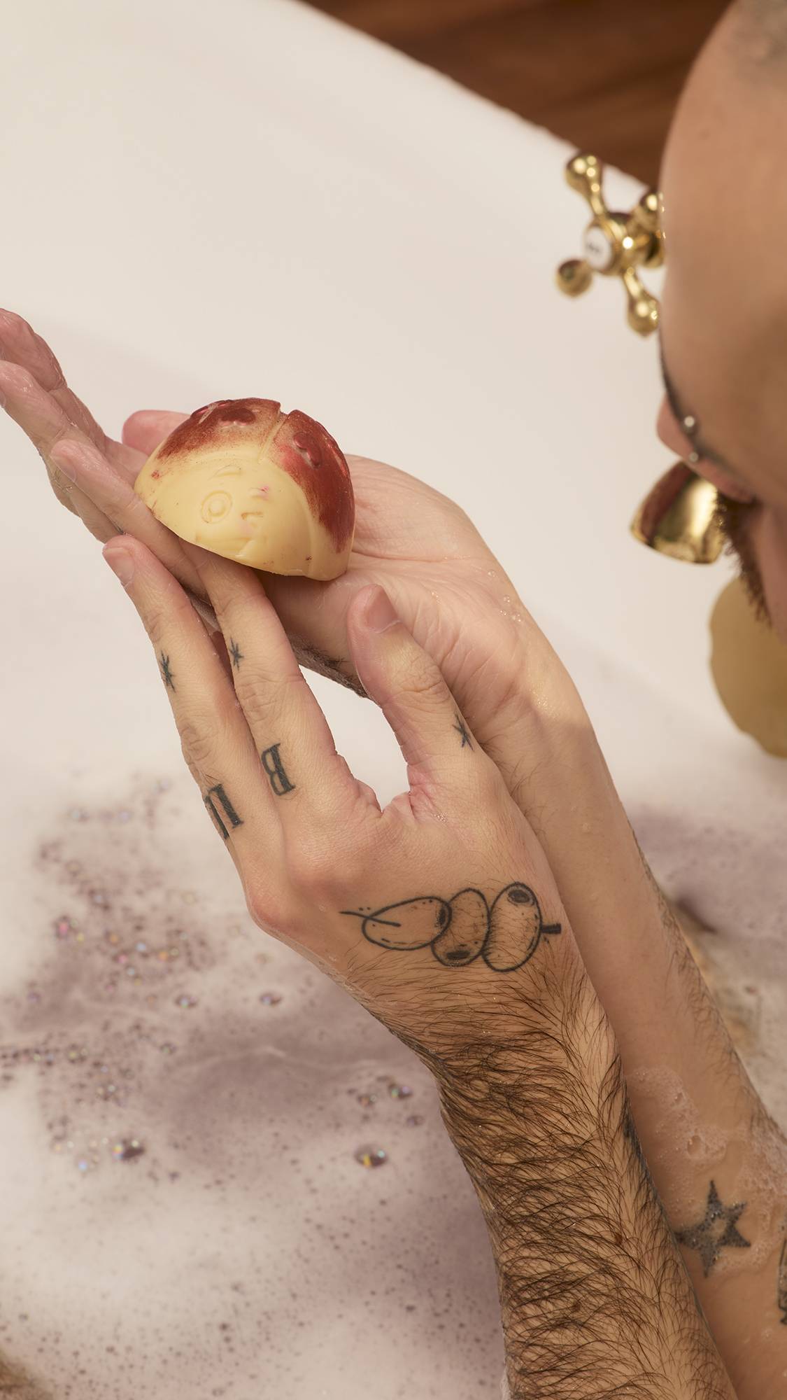 A person with tattooed hands holds a cream-coloured, ladybird-shaped Love Bug Scrubee solid body scrub over a tub of bubbly water. The bath bomb has red-shimmering wings and a winking face, shown as it is held over the bath.