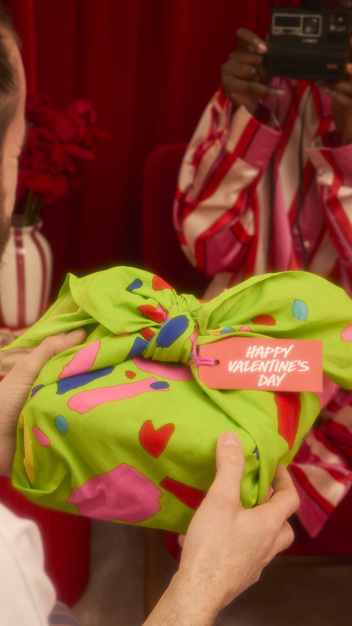 Hands hold a gift wrapped in the green Love Knot Knot Wrap, featuring abstract shapes and a red tag reading Happy Valentine's Day. In the background, a person in striped pajamas holds a camera to take a photo.