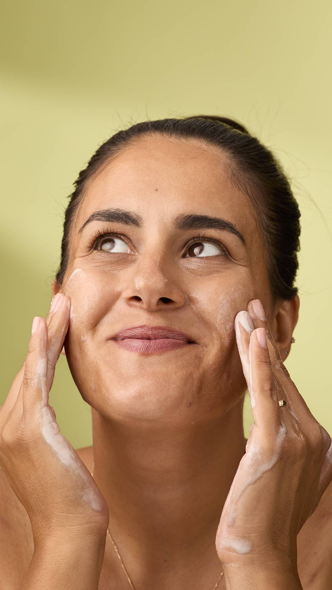 A smiling woman looks upwards as she massages the white lather from the Lucky Cat facial gummy into her cheeks. Her soapy hands frame her face against a plain, light olive-green background. She is wearing a delicate chain necklace.