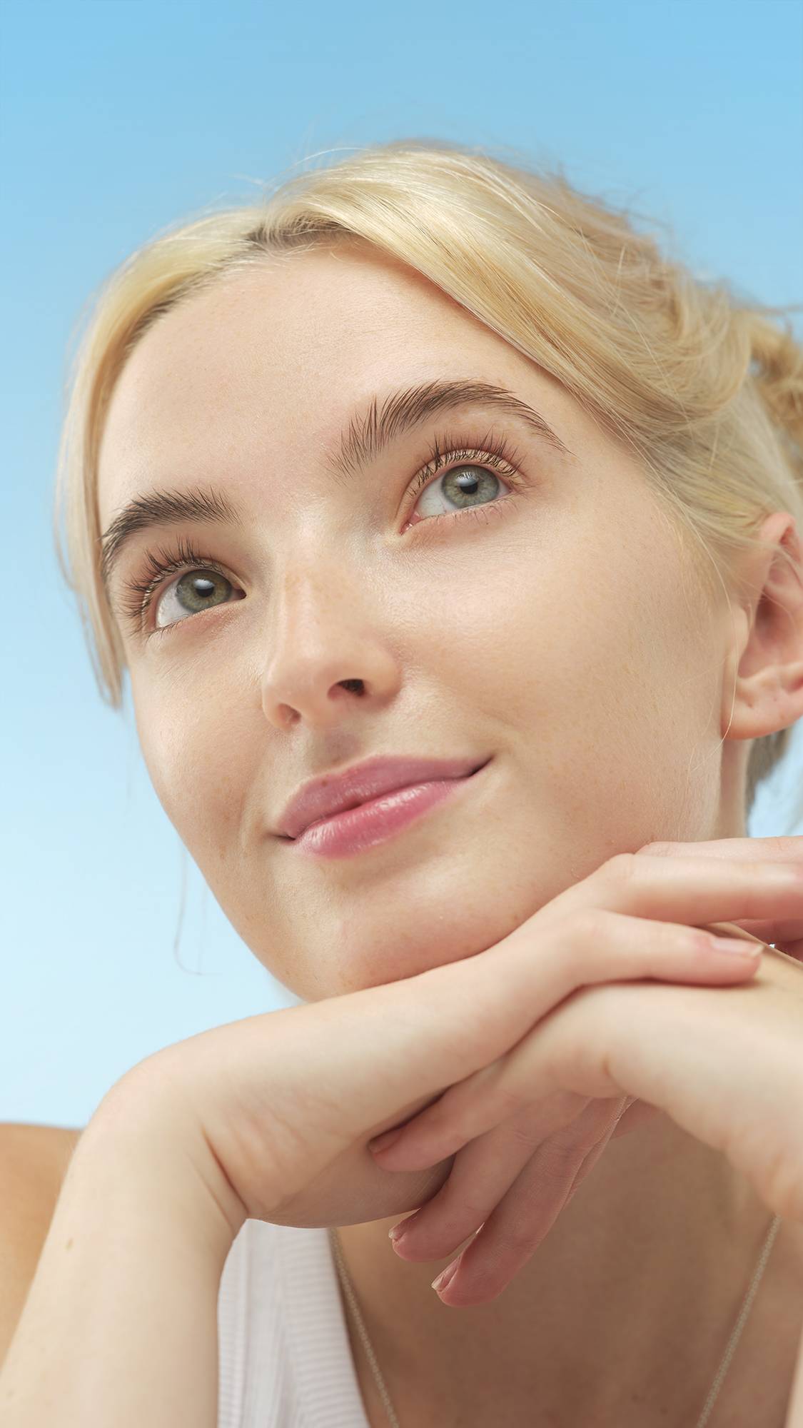 A close-up portrait of a smiling person with fair skin, blonde hair, and light eyes, looking up. Their hands cup their chin, and their face glows from the Luminator - skin tint, set against a bright blue background.