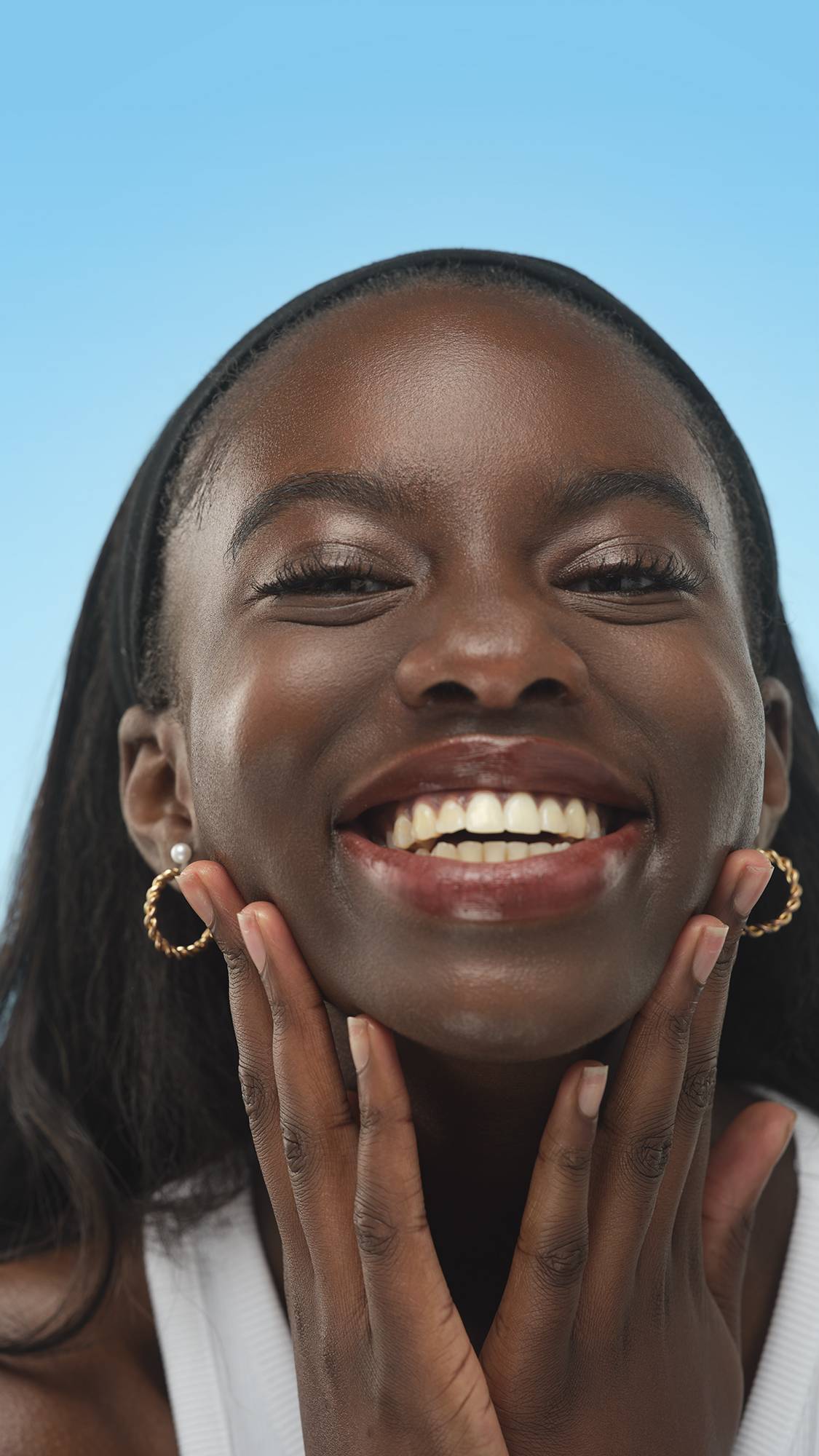 A person with dark skin and a black headband smiles widely, framing their face. They have long, defined lashes, likely from Lush Lashes ~ Mascara, and wear gold and pearl earrings, against a blue background.