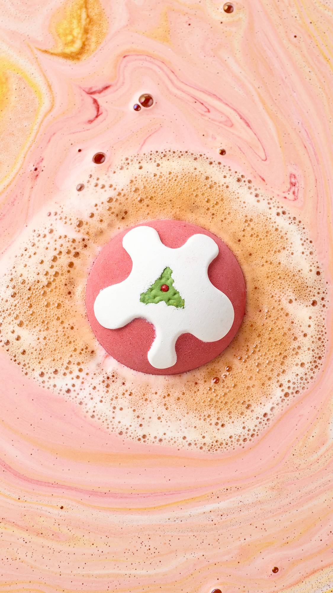 A Christmas pudding-shaped bath bomb floats at the centre of bathwater, releasing a ring of rich, creamy beige foam into pastel pink and soft orange marbled water.