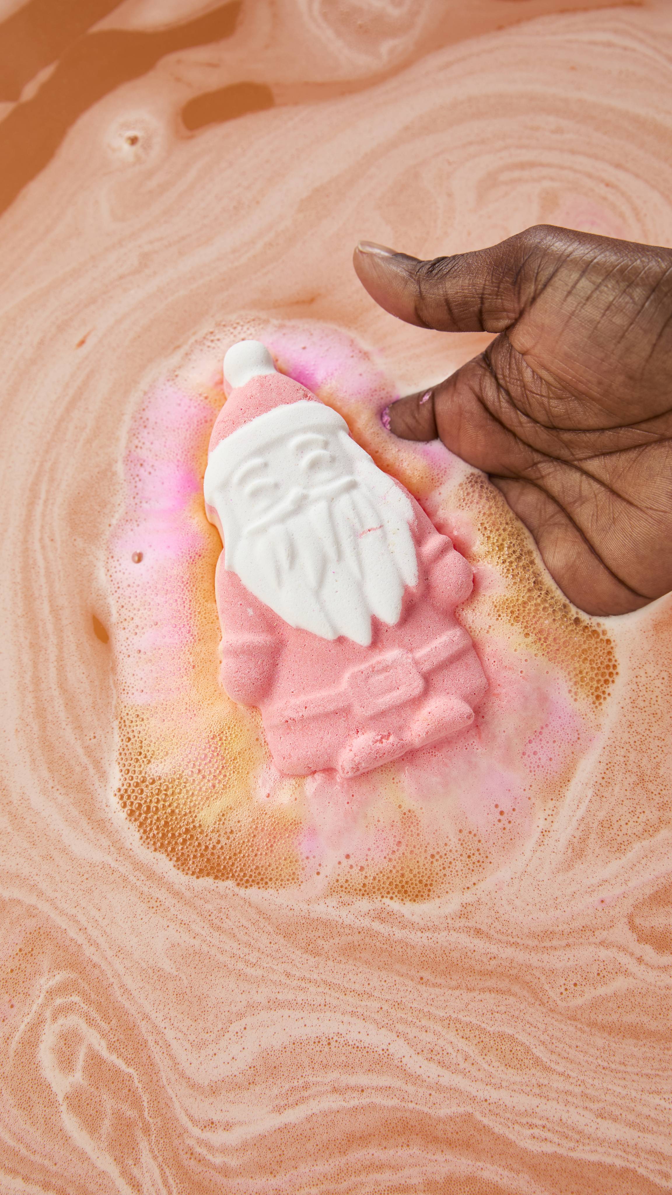 A pink Lush "Magical Santa" bath bomb fizzes into soft pink and golden foam as a hand holds it gently, just resting in the swirling peachy-pink bathwater.