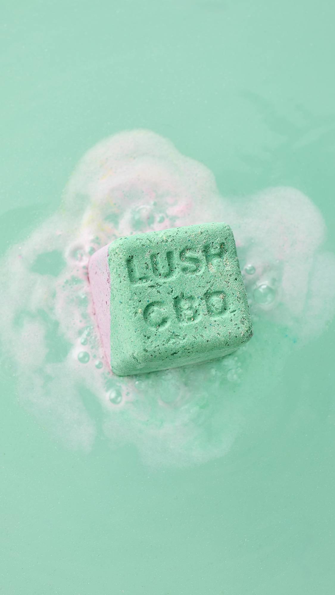 The square Magik bath bomb floats in mint green water, fizzing with a halo of white foam. The green face, embossed with "LUSH CBD," is visible as a hint of pink from its other side bleeds into the surrounding bubbles.