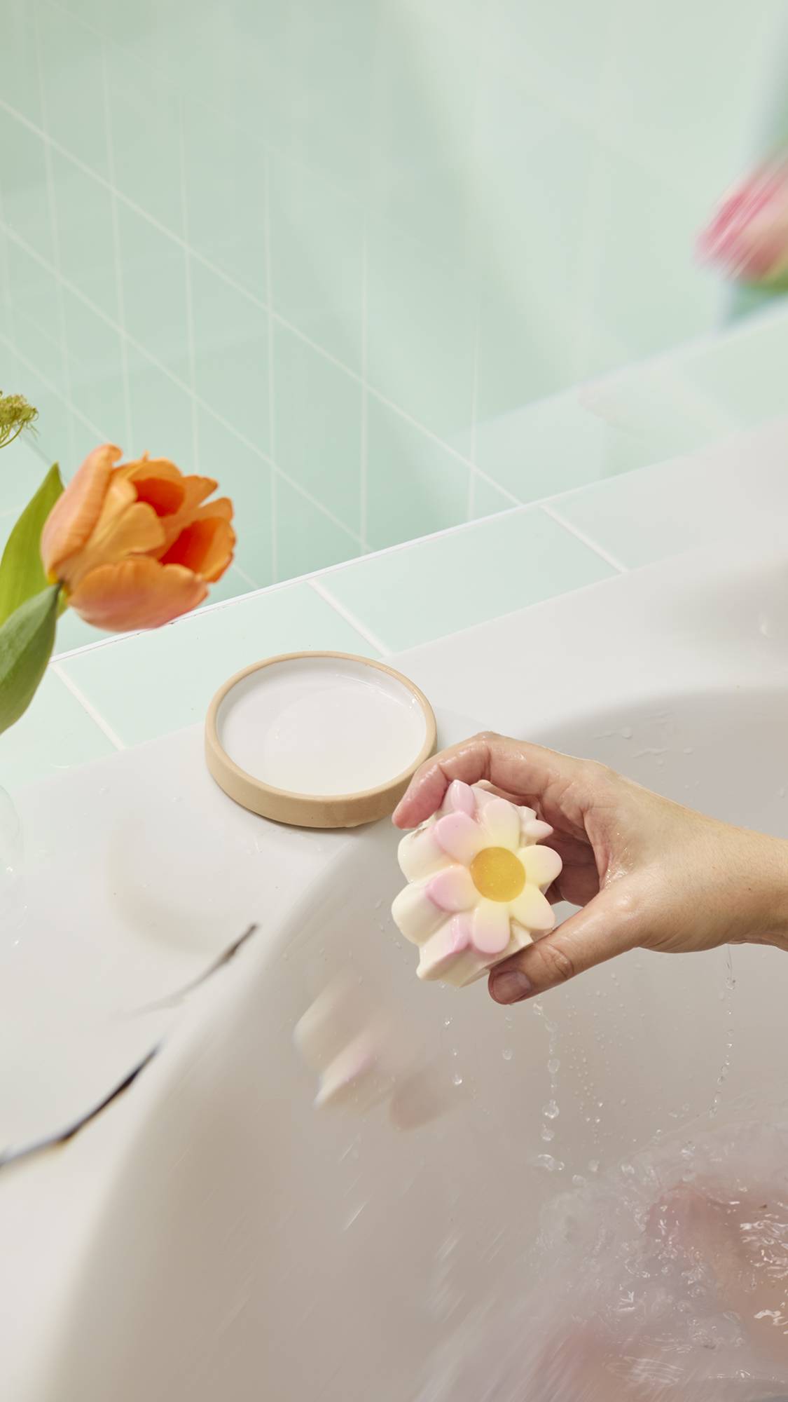 A hand holds a wet, daisy-shaped "Making Daisy Chains" bar over a bath, dripping water. The pink-tipped petals are framed by a blurred orange tulip and green tiles.
