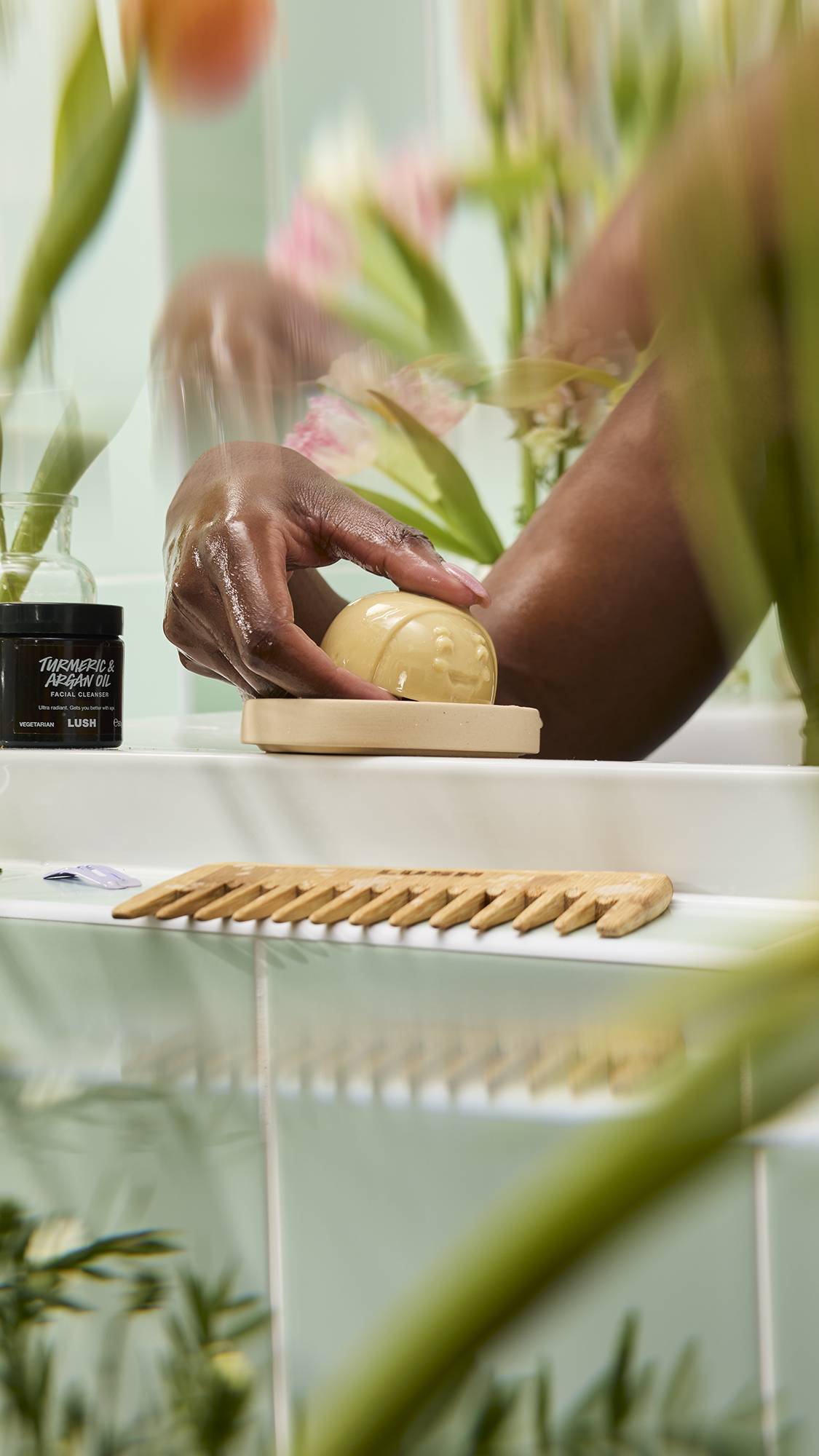 A hand reaches for a bee-shaped "Mama-chino Scrubee" bar on a dish. A "Turmeric & Argan Oil" cleanser and wooden comb sit nearby against mint green tiles and flowers.