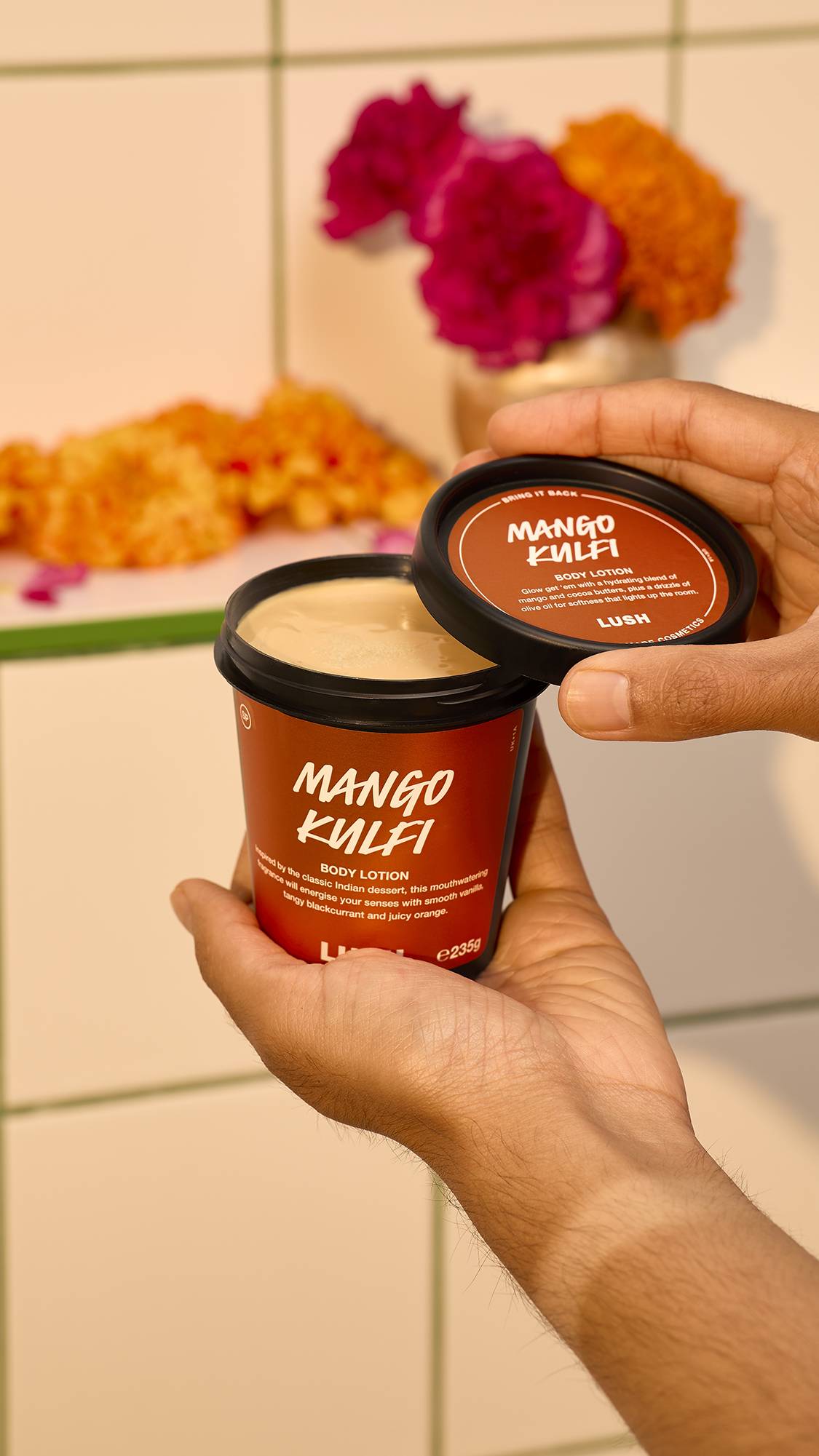 A pair of hands holds open a black tub of Lush Mango Kulfi body lotion to reveal the smooth, light yellow cream inside, against a tiled bathroom background with colourful marigold flowers.