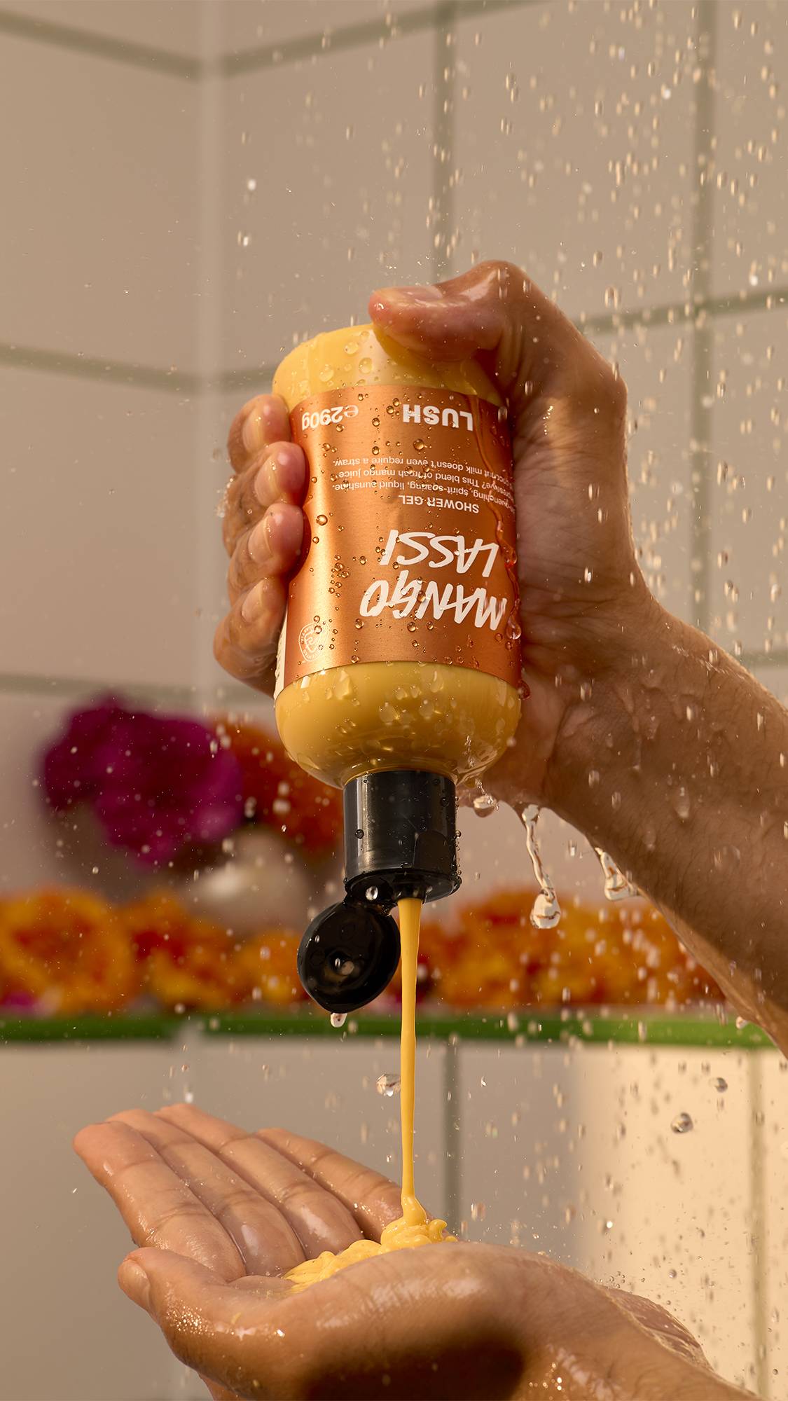 A hand squeezes yellow Lush Mango Lassi shower gel upside down into another open hand under falling shower water, against a tiled wall backdrop with blurred flowers.