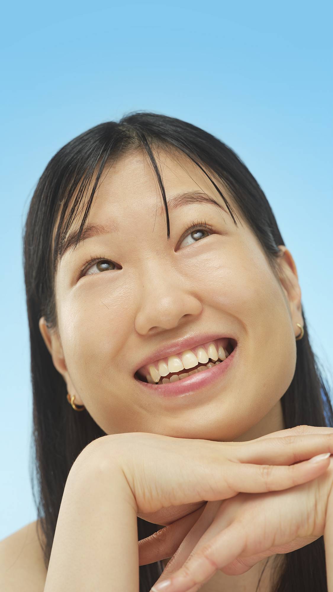 A person with long dark hair and Medium Cool Skinwear foundation applied is smiling widely while looking up. They rest their chin on their stacked hands against a light blue background.