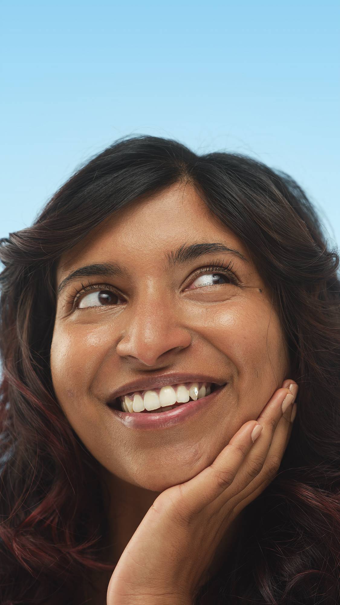 A smiling person with Medium Dark Neutral Skinwear foundation applied, rests a hand on their chin and looks up and to the side against a light blue background.