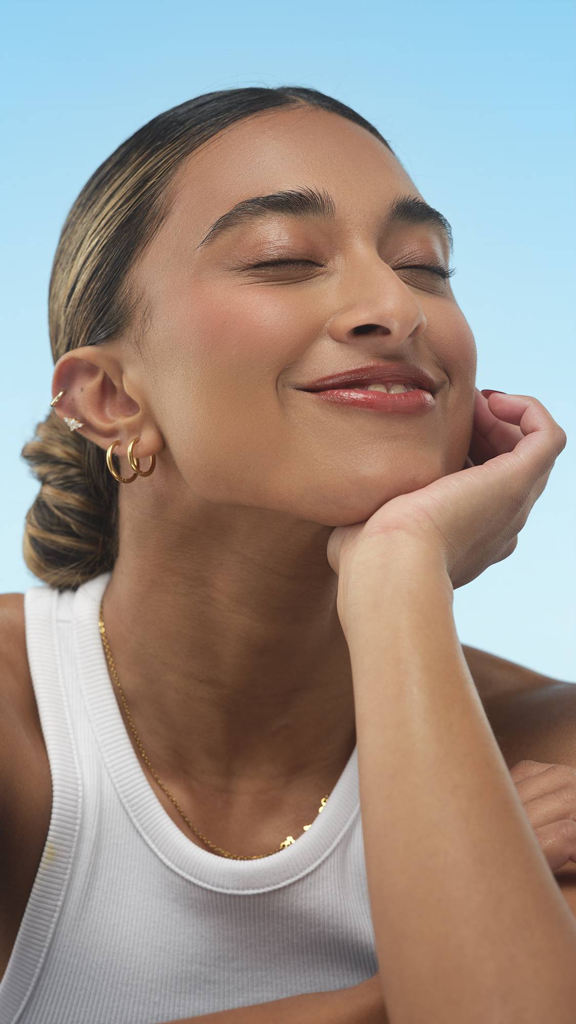 A close-up vertical portrait of a person with medium-dark skin, smiling with eyes closed and chin resting on their hand. Their face has Medium Dark Warm Beauty Balm on, and they wear gold jewelry, set against a bright blue background.