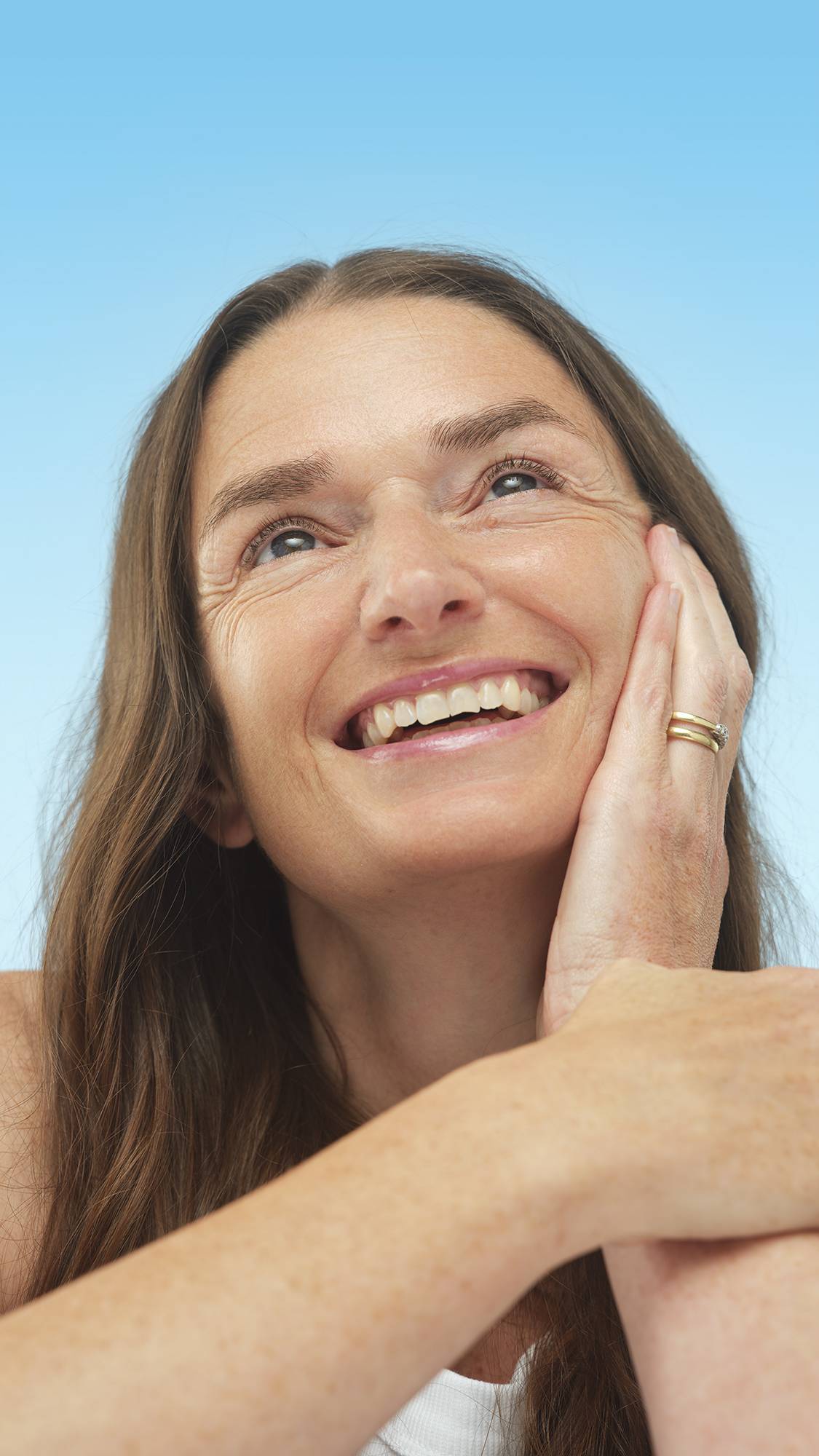 A smiling person with long brown hair and Medium Neutral Skinwear foundation applied, looks up while resting a hand on their cheek. They are wearing a simple gold ring against a light blue background.