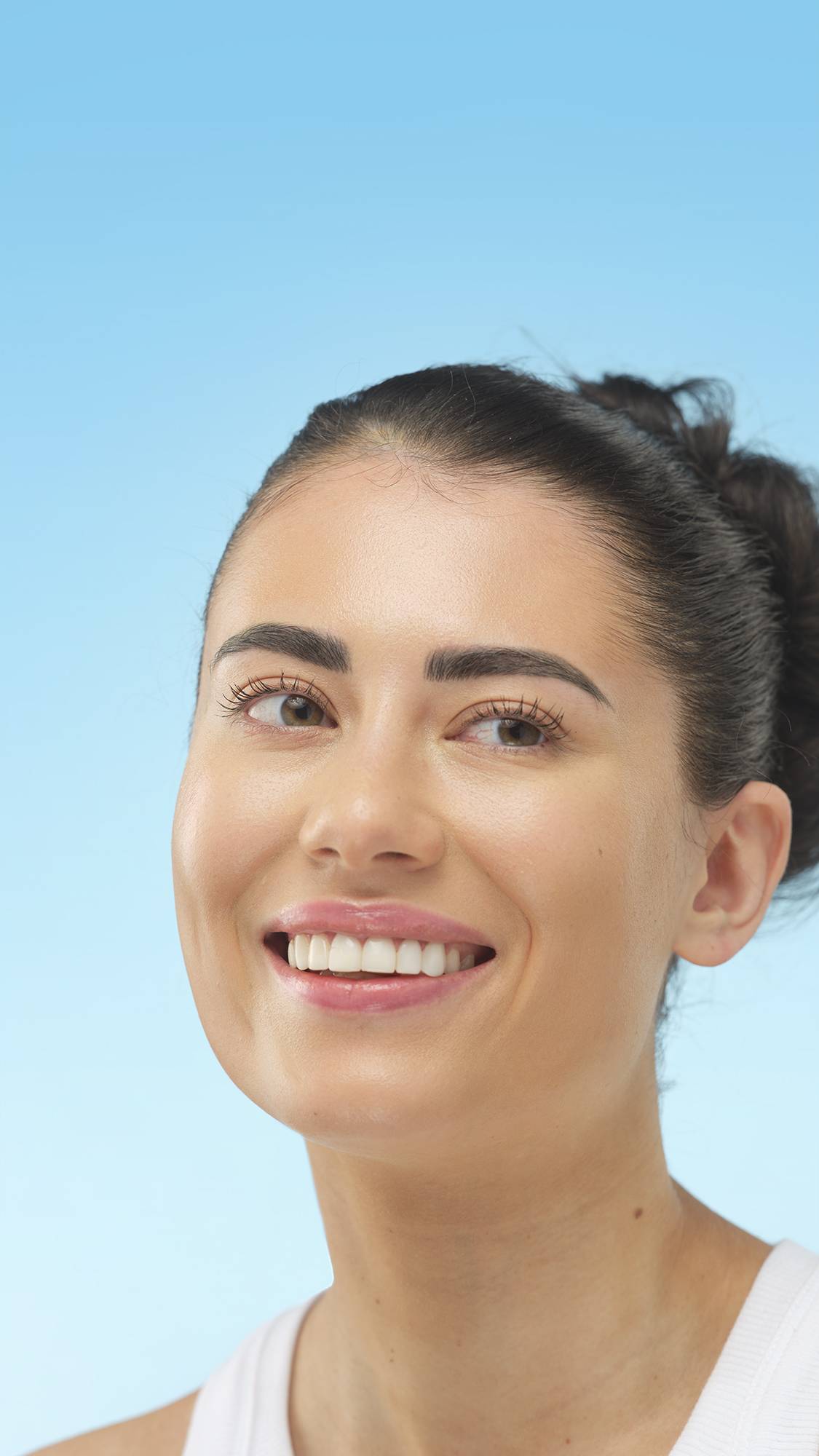 A smiling person with an updo and bold eyebrows looks to the side. They have Medium Warm Skinwear foundation applied and are posing against a light blue background.