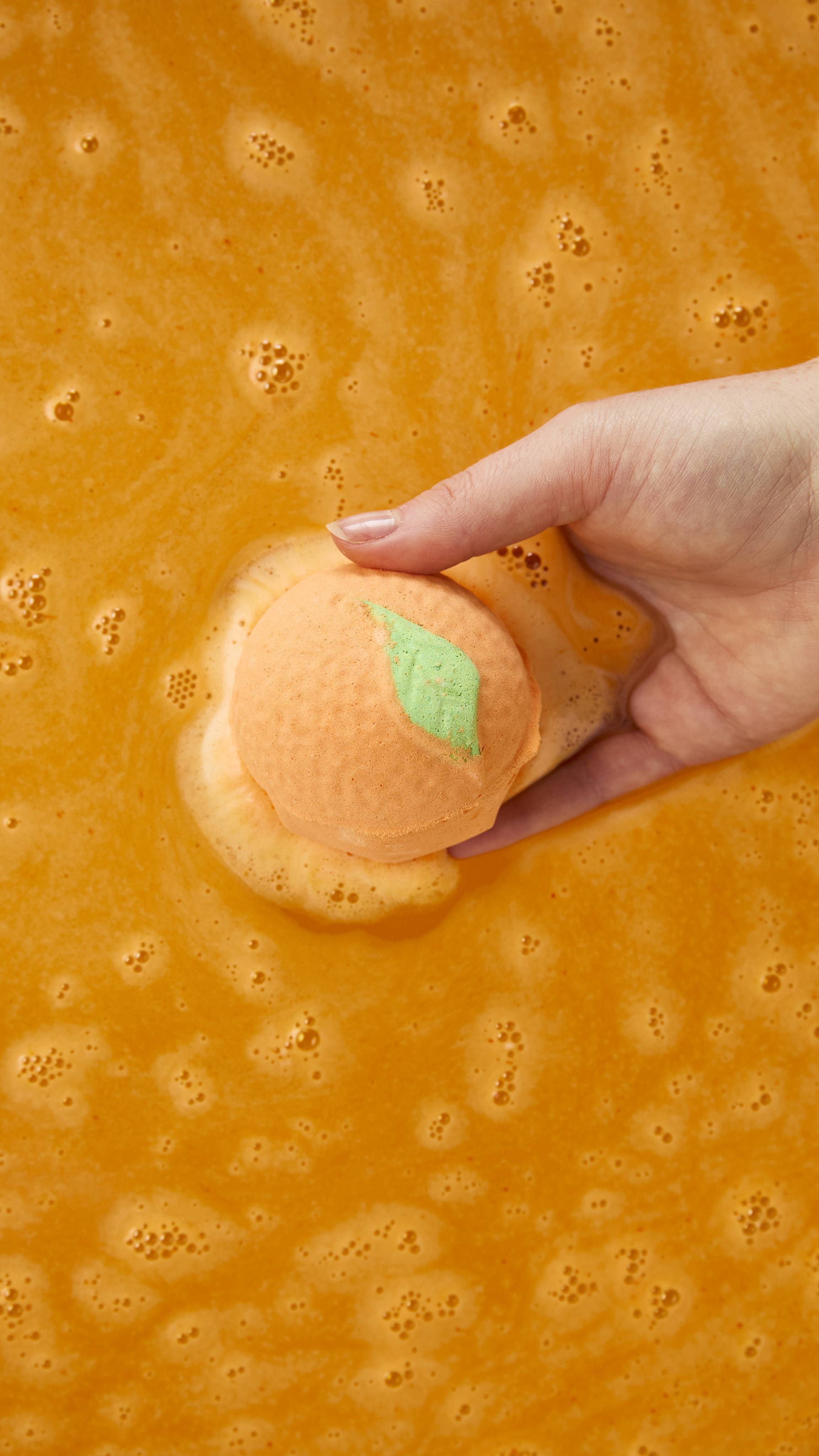 An orange-shaped bath bomb with a green leaf detail fizzes into rich, creamy foam as a hand dips it into bright orange bathwater.