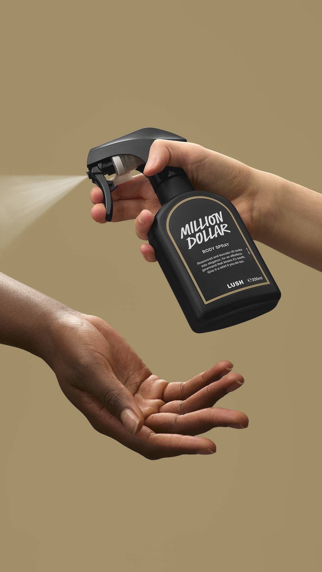 A person's hand presses the nozzle of a black Lush 'Million Dollar' body spray bottle, releasing a fine mist onto another person's open palm. The action is set against a solid, plain beige background. 