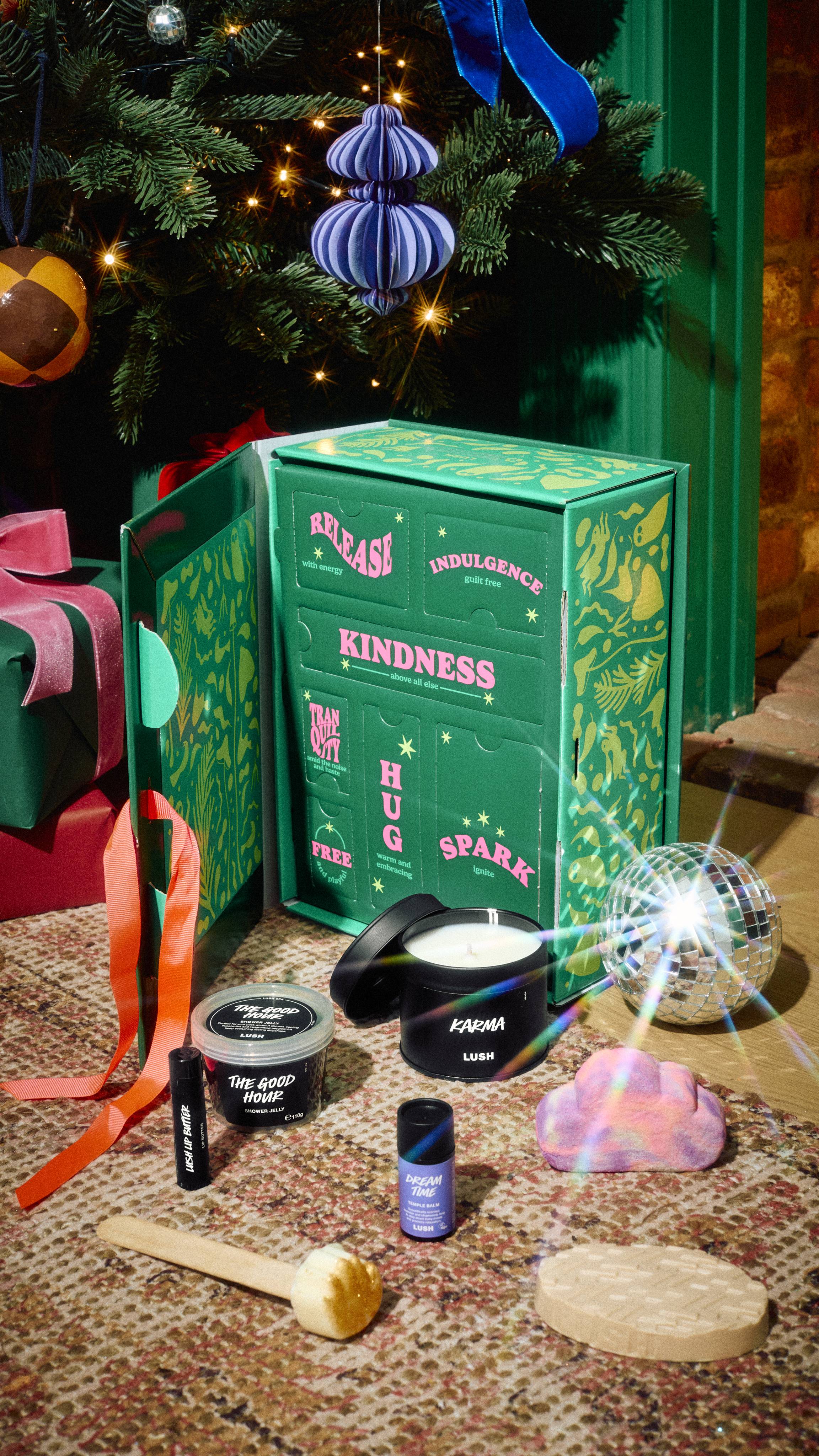 An open Lush Mindful Moments green gift box with labelled doors, displayed on a rug with a lit Karma candle, Dream Time temple balm, shower jelly, bubble bars, lip butter, and massage bar, beside a Christmas tree.