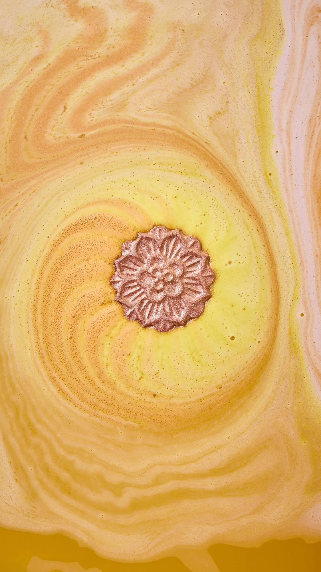 An overhead shot of a mooncake-shaped bath bomb dissolving in water. The bronze, floral-patterned product sits at the centre of a swirling vortex of warm yellow and creamy orange foam, creating a marbled effect across the surface.