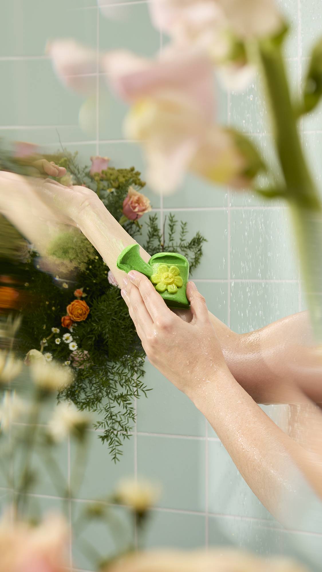 A person washes their arm with the green, watering can-shaped "Mum's Garden" soap. Mint green tiles and blurred flowers frame the shower scene.