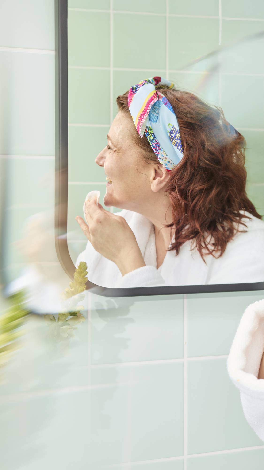 The image shows a model with wavy auburn hair in a bathroom mirror, smiling while cleansing her face with a cotton pad. She has fashioned the colourful "My Family Houses" fabric knot wrap into a stylish headband.