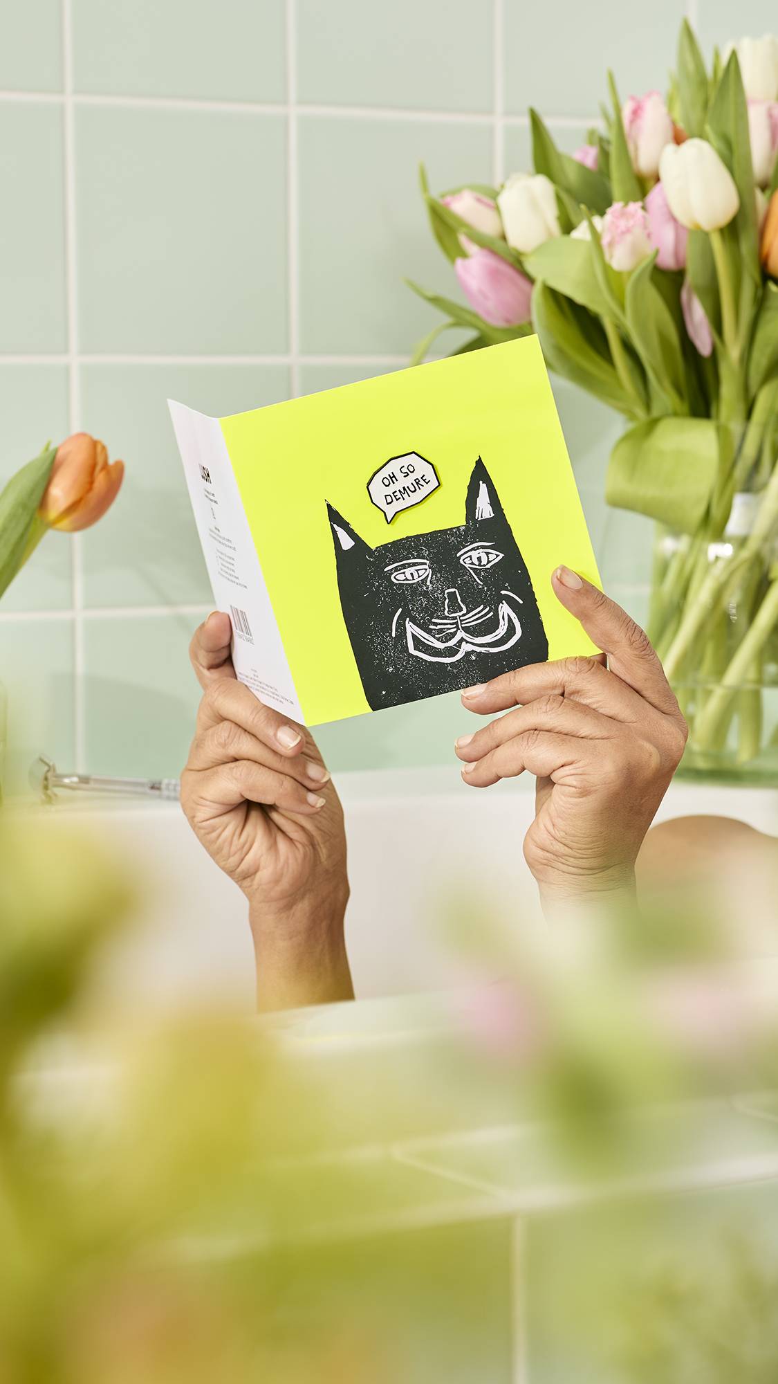 The image shows the model's hands over the edge of a bath, holding a green Oh So Demure card featuring a black cat illustration and a speech bubble reading ‘OH SO DEMURE’. The background shows pale green tiles and a bouquet of pink and white tulips.
