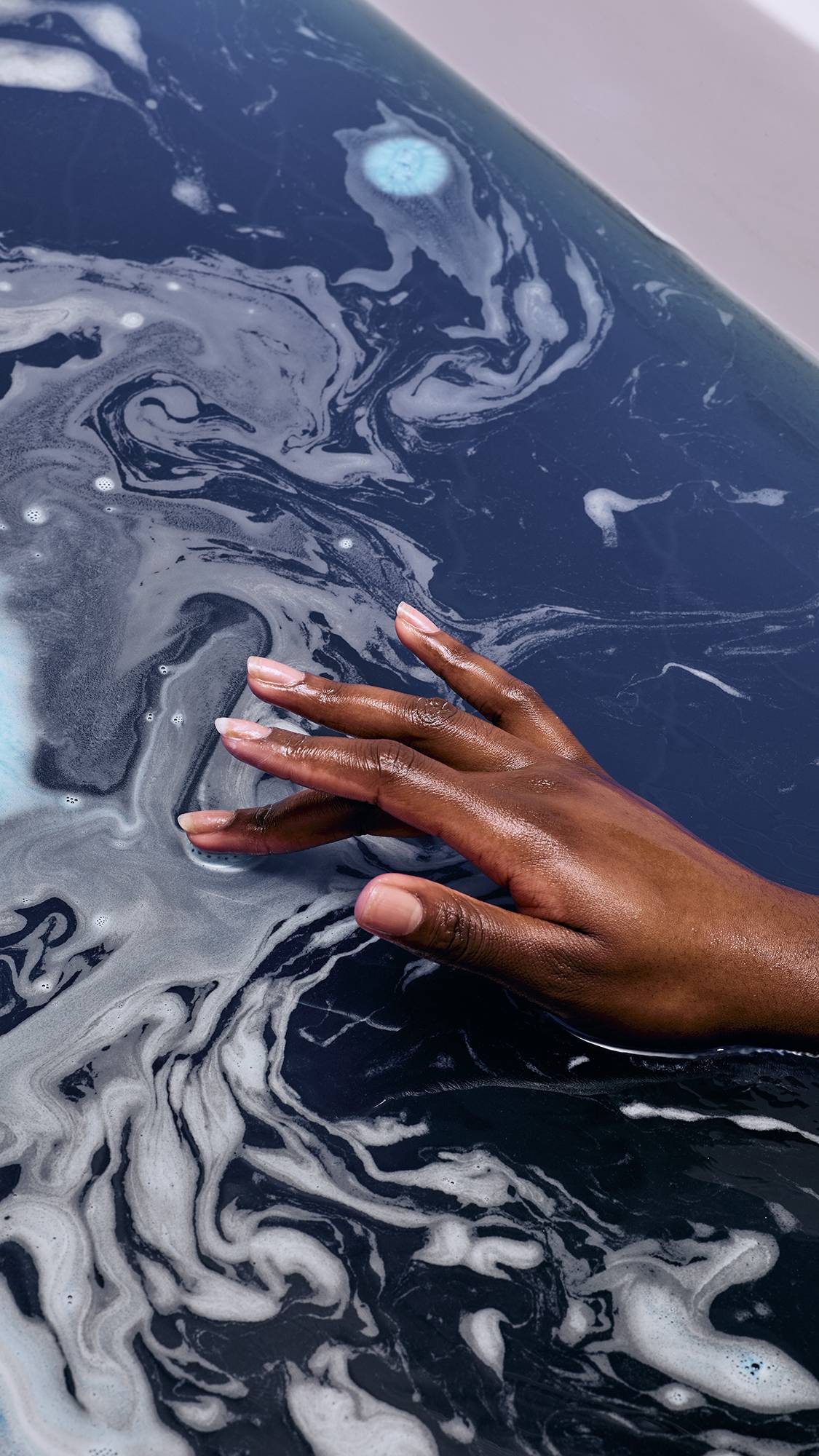 A model's hand with deep-toned skin rests on the surface of dark blue bathwater. Swirls of white and light blue foam create an ethereal, marbled pattern across the water, created by the "Pease" bath bomb.