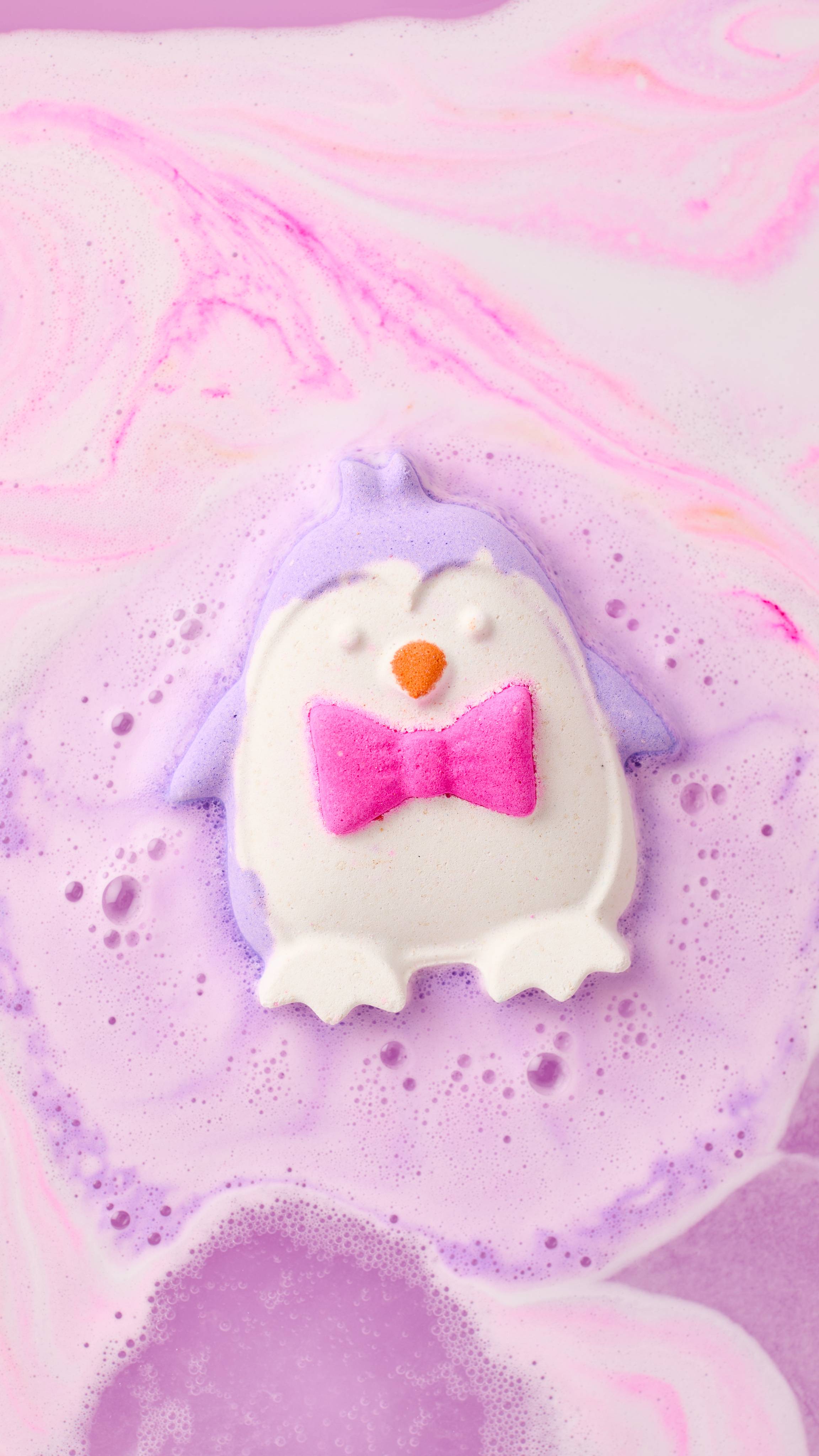 A lilac and white penguin bath bomb with a bright pink bow tie dissolves in bathwater, releasing a ring of fine purple foam across swirling pastel pink water.