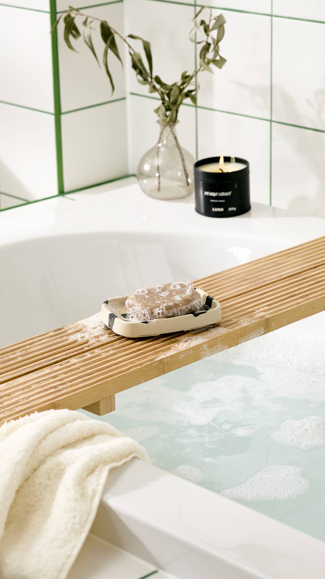 A bar of bubbly soap sits on a cow-print ceramic soap dish over a wooden bath caddy, resting across a tub filled with foamy bathwater. A black candle, vase, and white towel sit nearby against white tiled walls.