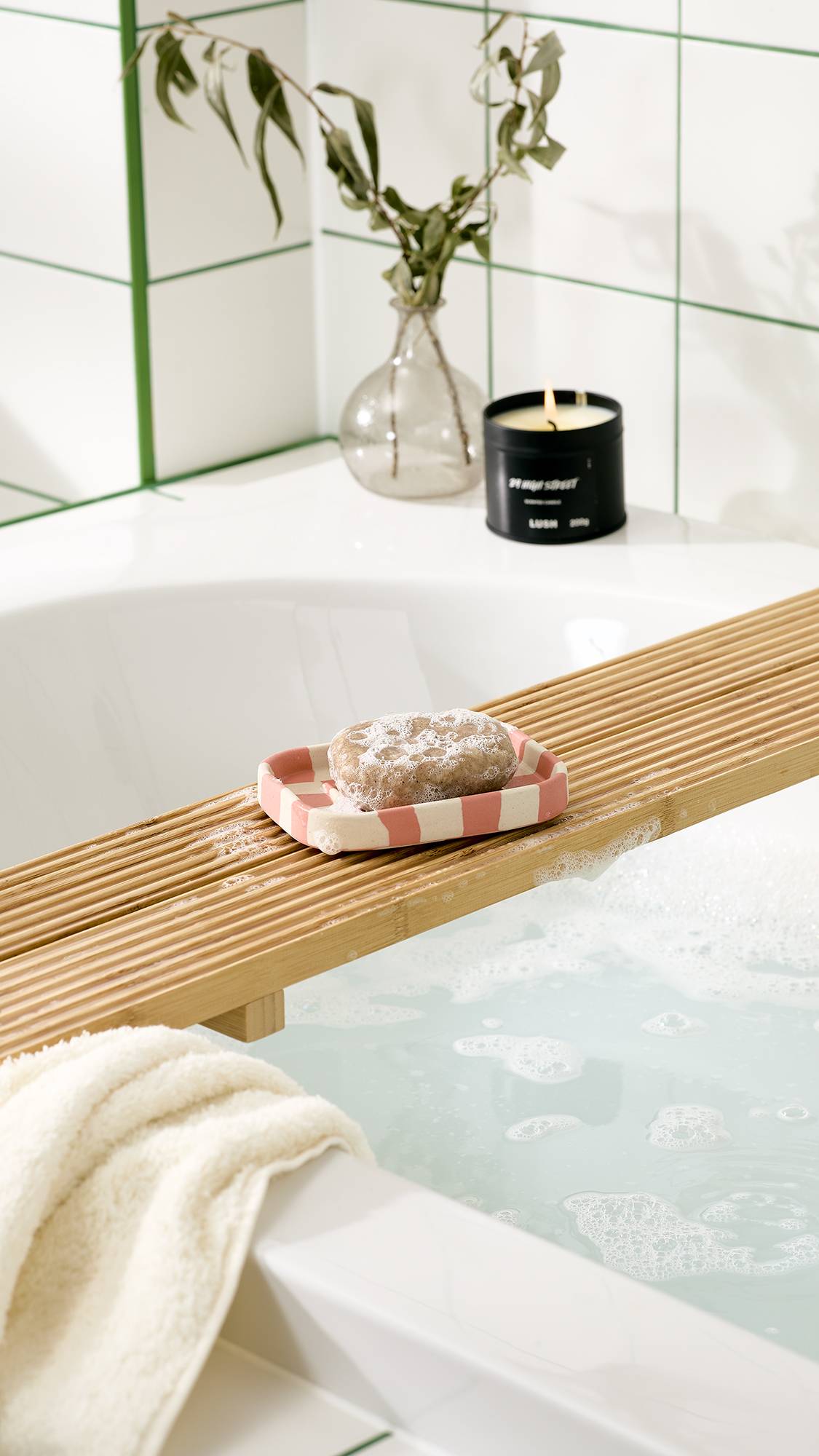 A bar of bubbly soap rests on a pink and cream chequered ceramic soap dish on a wooden bath caddy over a tub filled with foamy bathwater. A black candle, vase, and white towel sit nearby against white tiled walls.