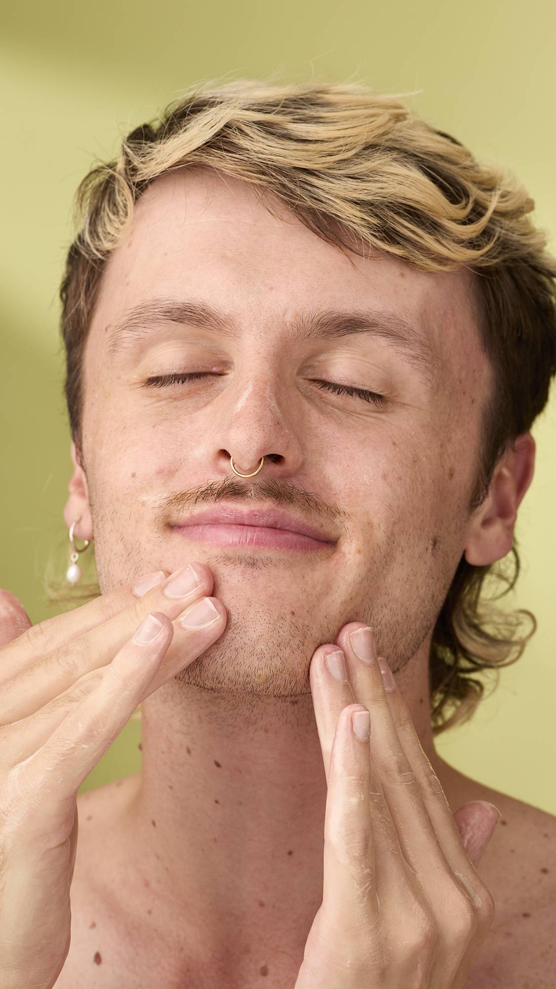 A close-up of a smiling person with blonde-highlighted hair and eyes closed, massaging their chin with white foam from the pumpkin facial gummy. They are cleansing their face against a plain, light olive-green background.