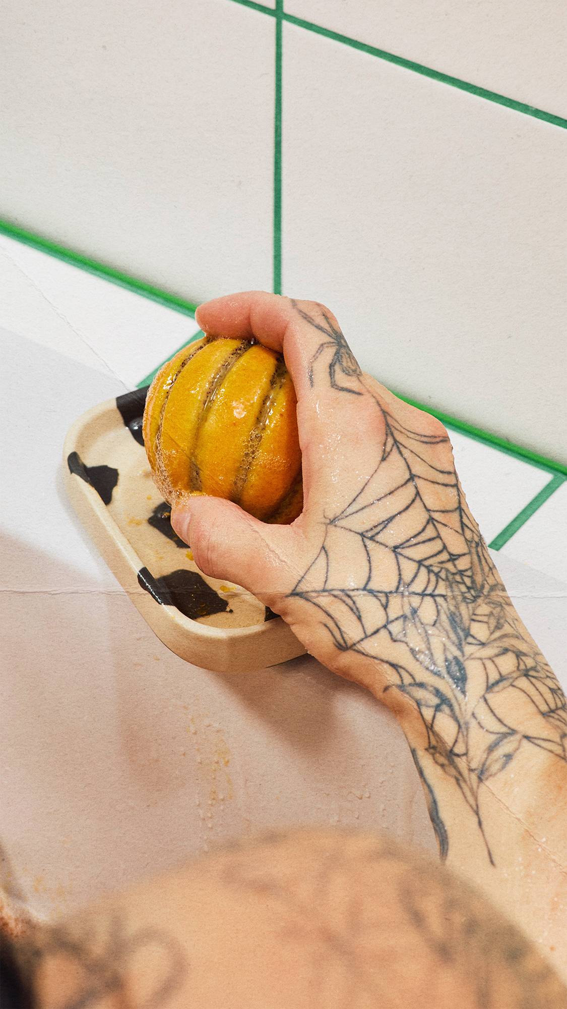 A tattooed hand with a spiderweb design picks up a small, orange pumpkin-shaped solid body scrub from a spotted soap dish beside a white tiled wall with green grout lines.