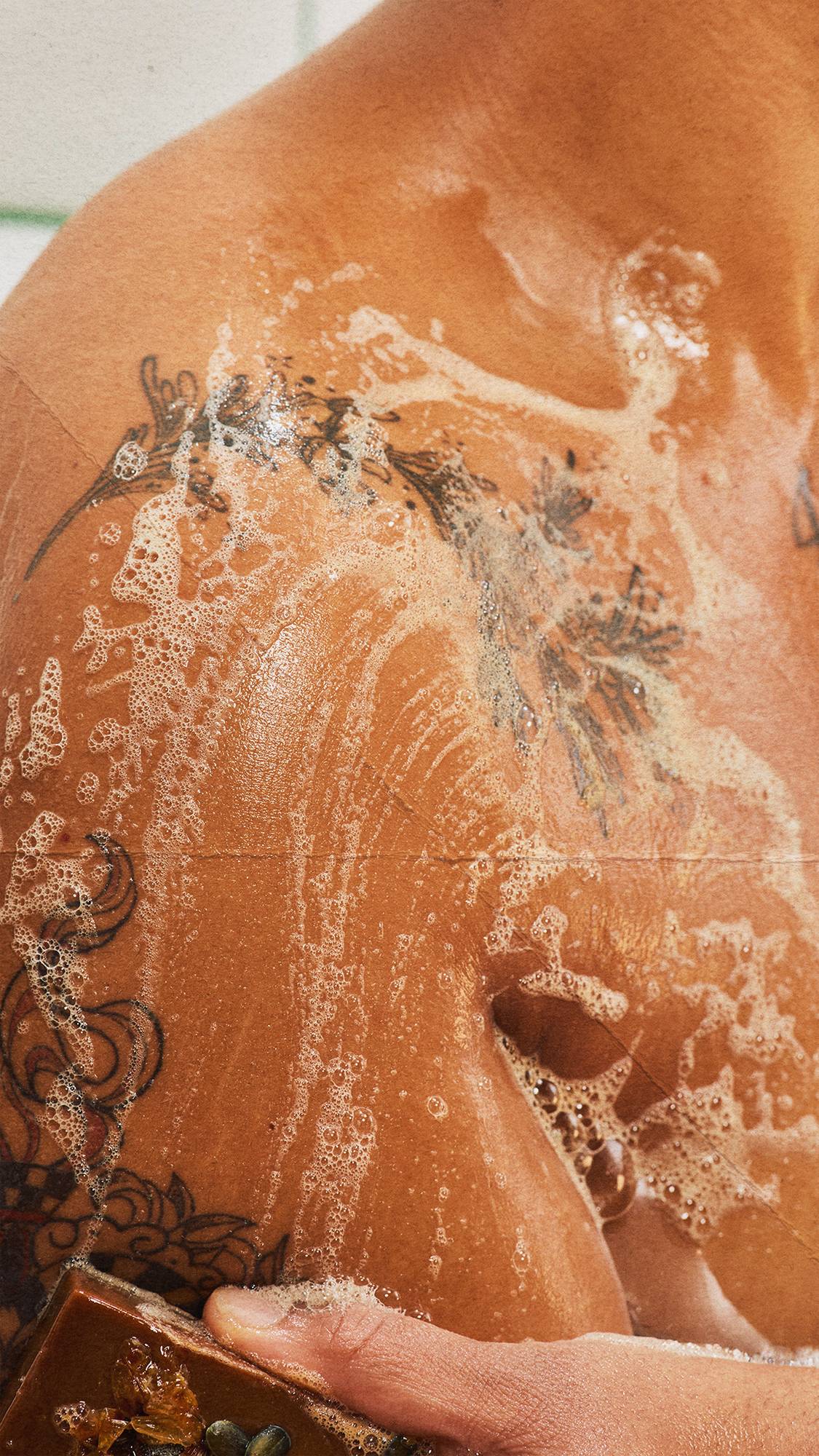 A close-up shot of a hand lathering a rectangular orange soap over wet, tattooed skin, creating rich white suds across the shoulder and chest.