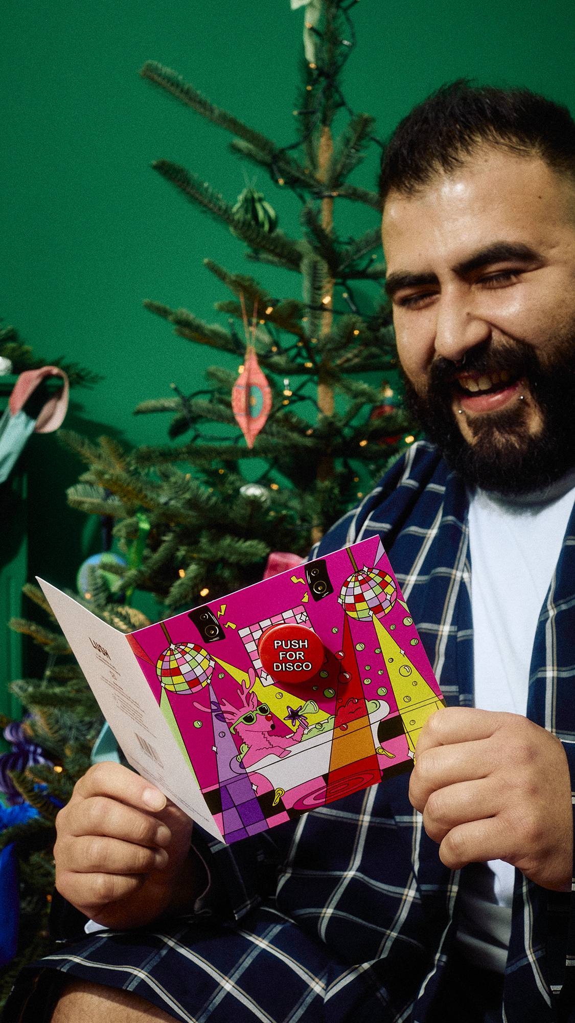 A smiling man with a beard wearing navy blue plaid pyjamas holds open a vibrant pink Lush Christmas card featuring a red "Push for Disco" badge, sitting in front of a decorated Christmas tree.