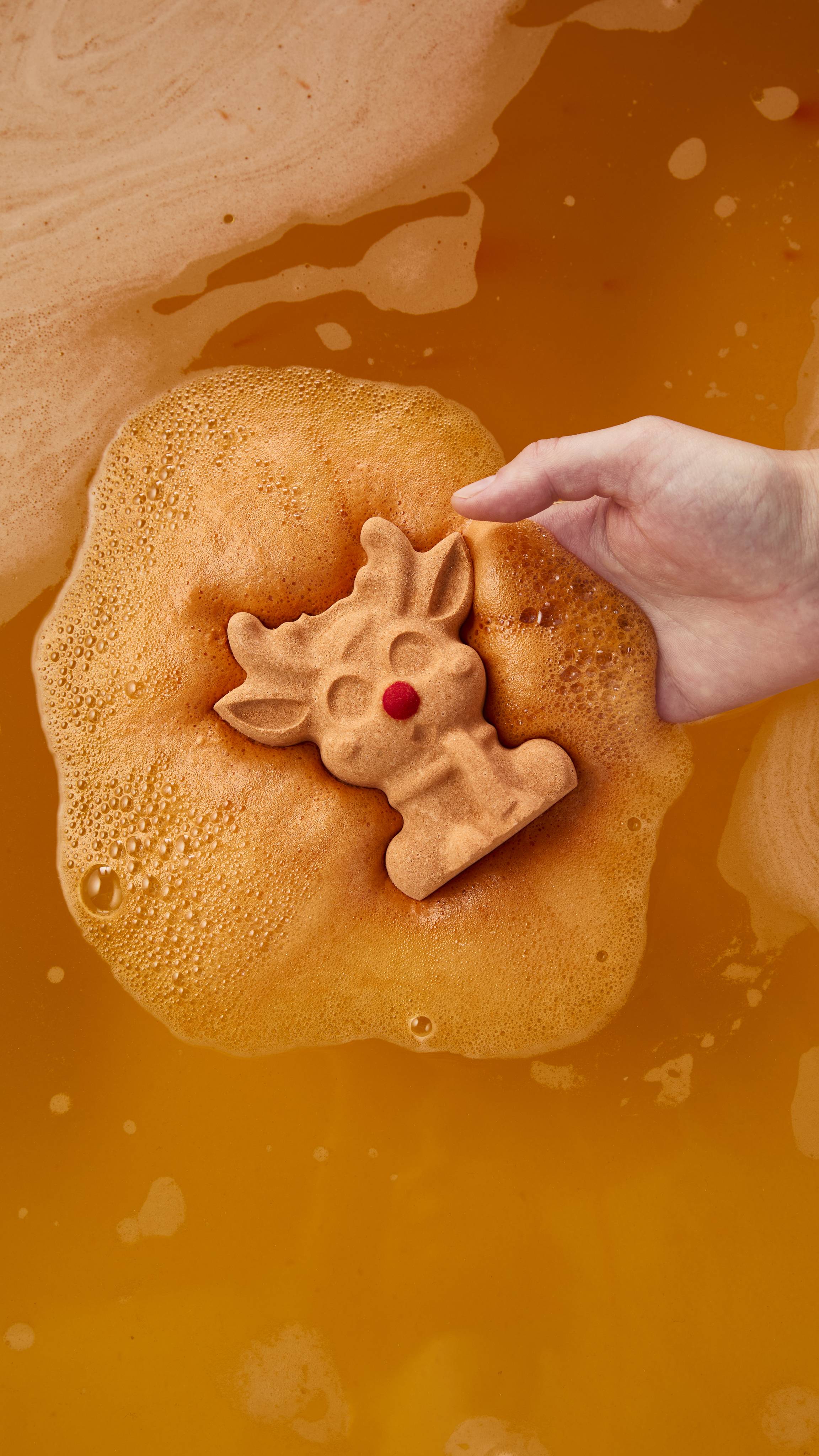 The image shows a hand gently placing a reindeer-shaped bath bomb on the surface of warm orange bathwater, fizzing into a thick layer of creamy golden foam