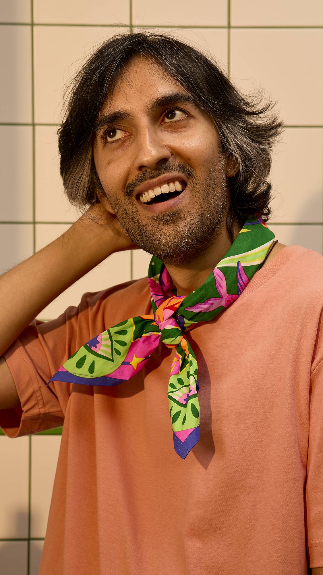 A model smiles while wearing a vibrant green, pink, and orange patterned Lush Knot Wrap tied around their neck, fashioned as a scarf, posing against a white tiled wall backdrop.