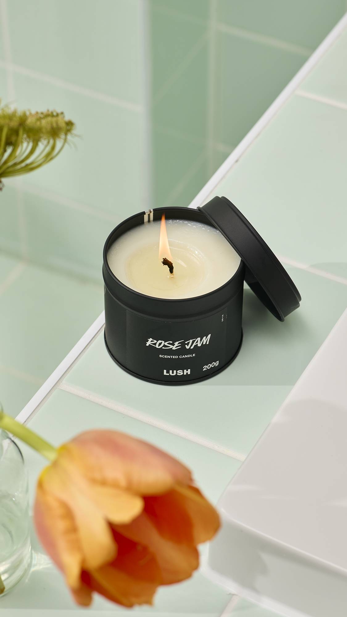 A close-up, high-angle image of a lit Lush "Rose Jam" scented candle in a round black tin. The flickering flame sits in a pool of melted white wax. It rests on a pale green tiled surface next to a blurred orange tulip.