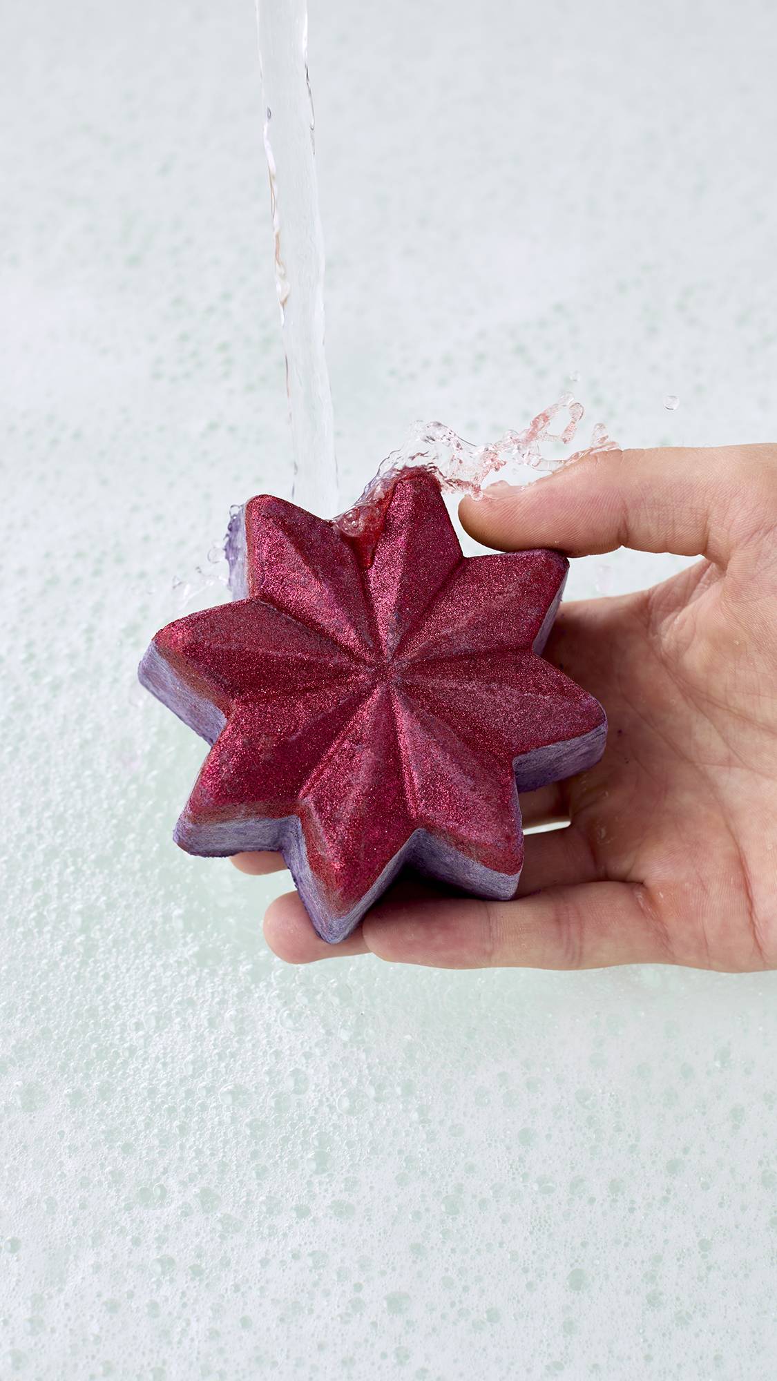 A hand holds a shimmering ruby-red and purple star-shaped bubble bar under running tap water over bathwater covered in soft white bubbles, splashing gently as it starts to dissolve.