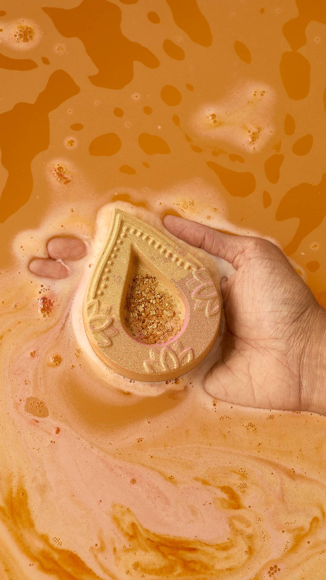 A hand gently supports a golden diya-shaped Lush "Saanjh" bath bomb floating on vibrant orange bathwater as light foam and shimmer begin to dissolve around it.