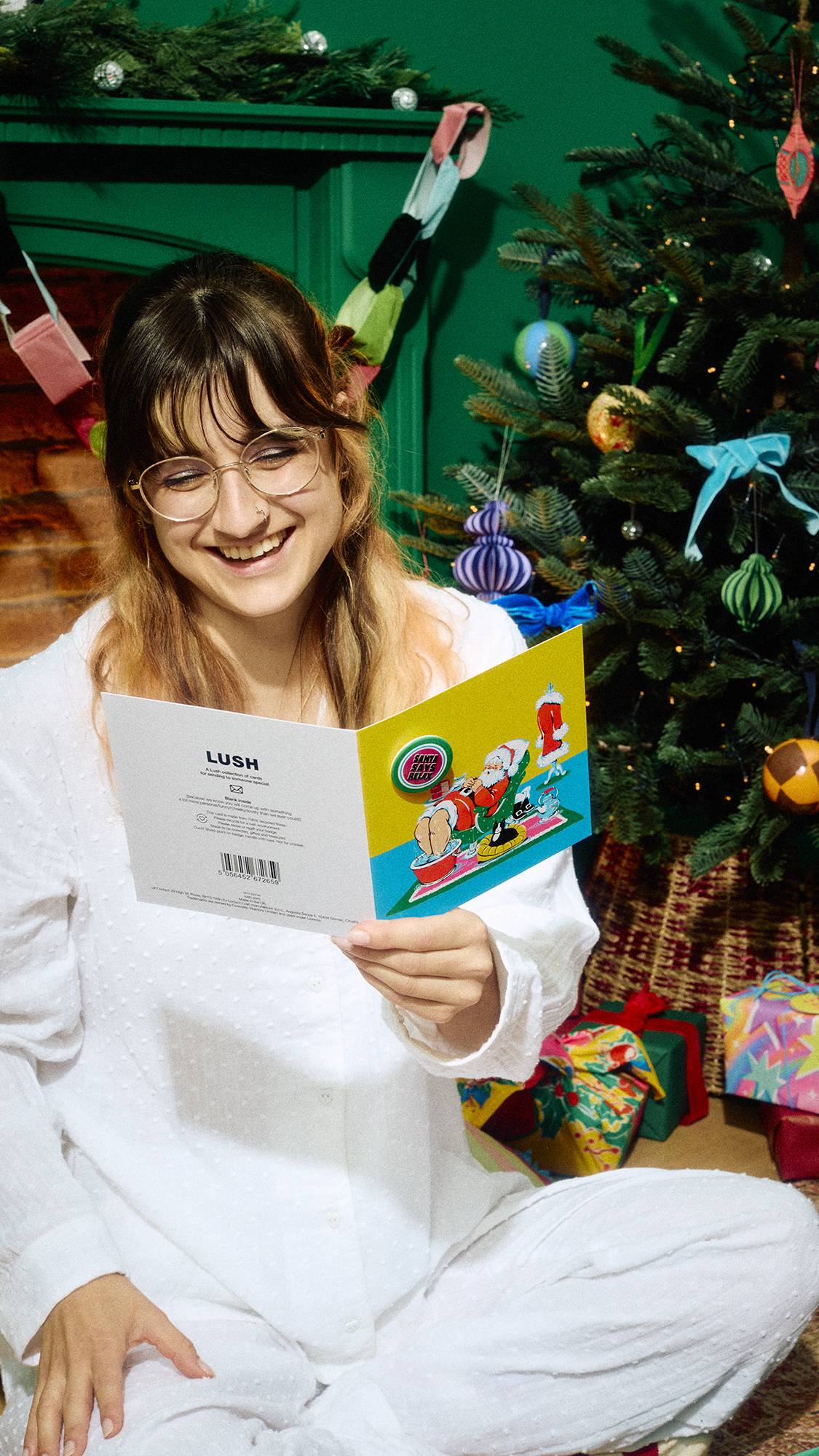 A smiling person in white pyjamas and glasses sits cross-legged holding open a yellow Lush Christmas card depicting a relaxing Santa, positioned next to a decorated Christmas tree and wrapped gifts.